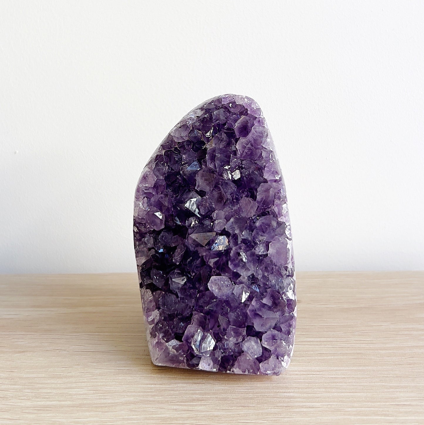 Close-up view of a sparkling amethyst crystal cluster, approximately 10cm tall, standing on a wooden surface against a minimalistic white background. | Simply Esoteric