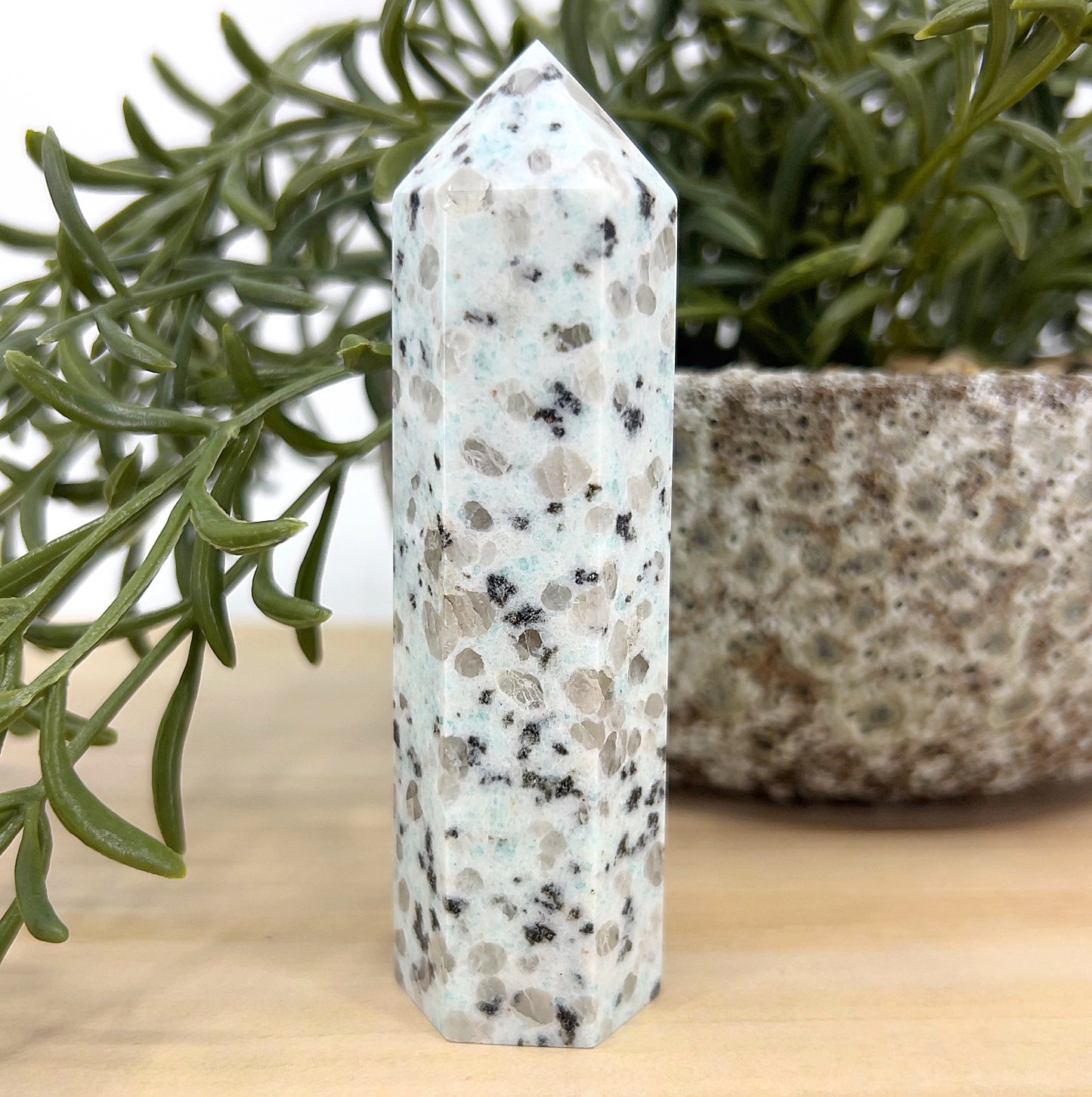 Kiwi Stone crystal tower approx. 10 cm tall with mint-green tones and black tourmaline speckles, displayed beside a green plant and stone pot – a healing crystal for calm energy and emotional balance.