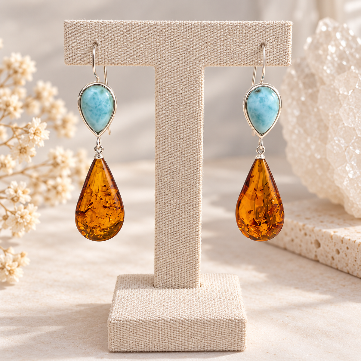 A pair of teardrop-shaped drop earrings displayed on a beige jewelry stand. Each earring features Larimar in a silver bezel at the top and a large amber teardrop drop below.