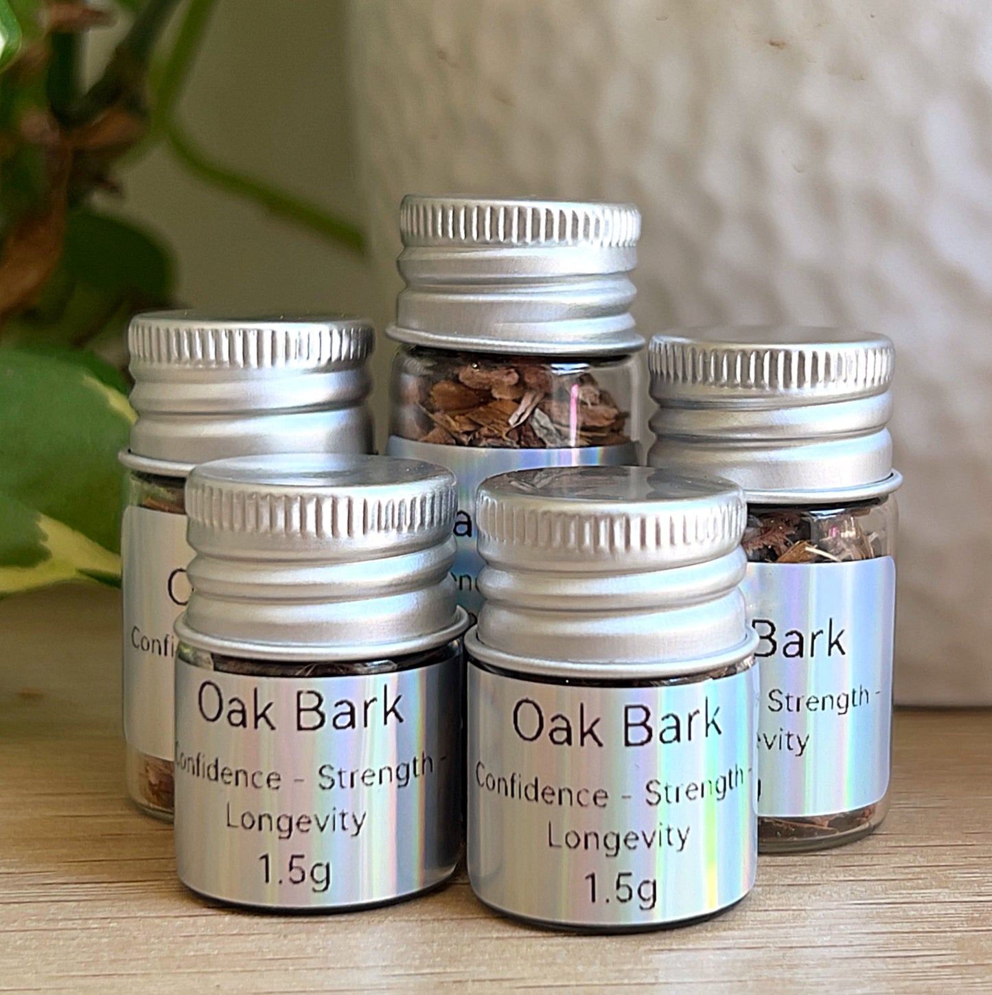 Small jars of oak bark (1.5 g, 2 g, and 3 g) with silver lids and labels, grouped on a wooden surface beside green plant leaves and a white pot.