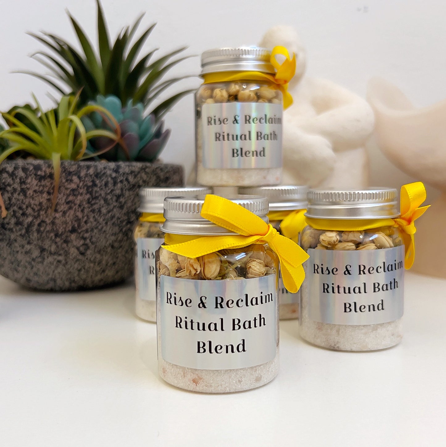 Jars of 'Rise & Reclaim Ritual Bath Blend' with yellow ribbons on a white surface.