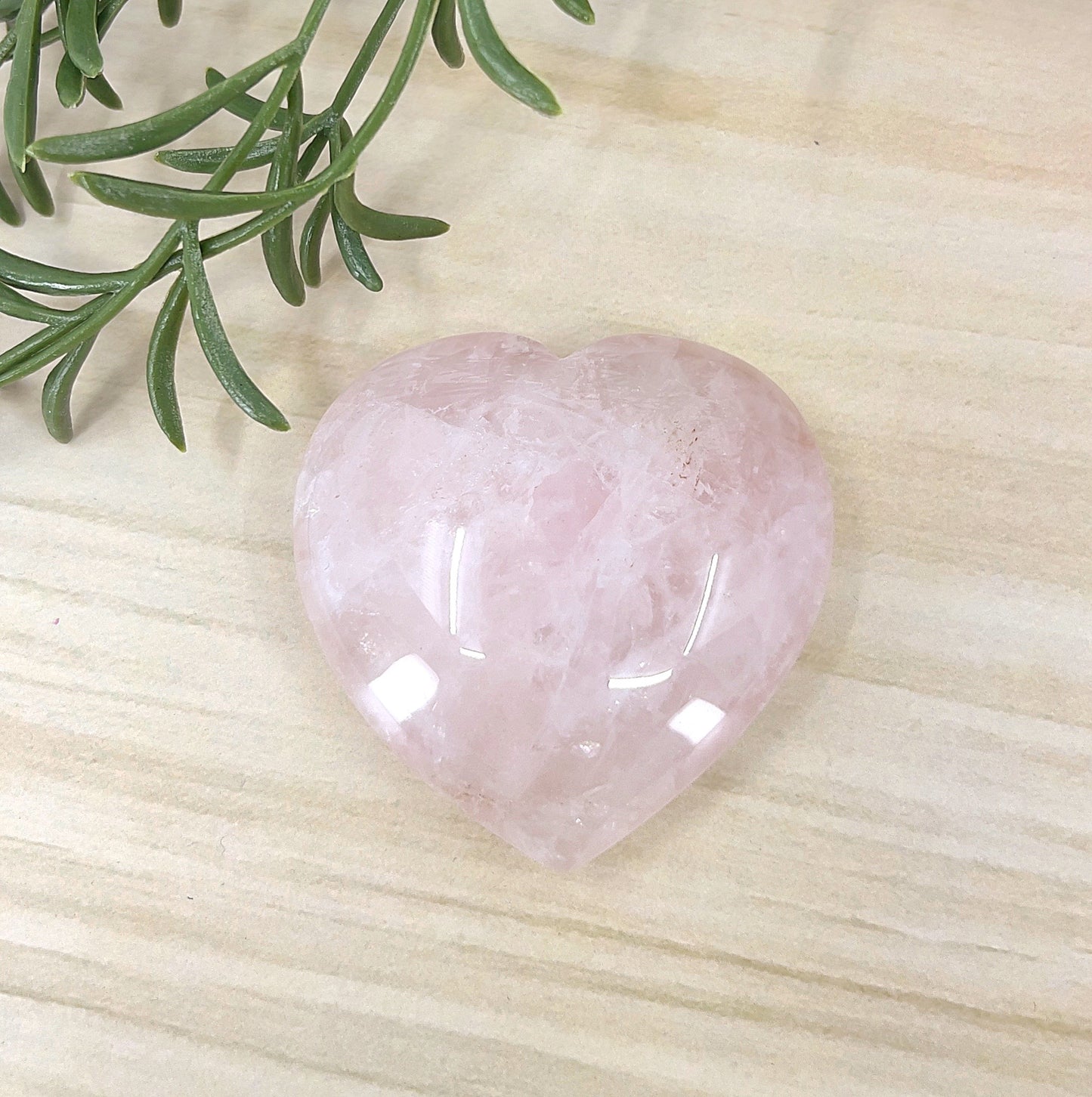 Rose Quartz puff heart crystal approx. 6 cm, polished soft pink stone on a light wooden surface beside greenery – symbolising love, emotional healing, and compassion.