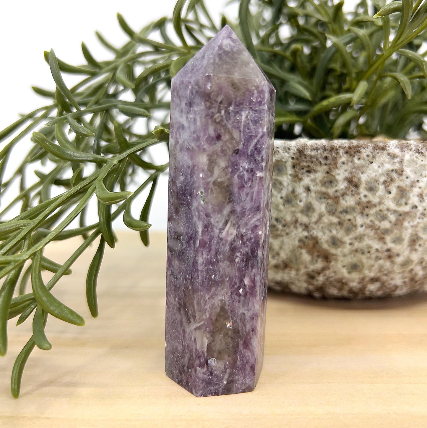 Unicorn Stone tower 9 cm tall showing soft lavender, pink and smoky tones, placed beside a green plant and stone pot – a calming crystal for emotional balance and healing.
