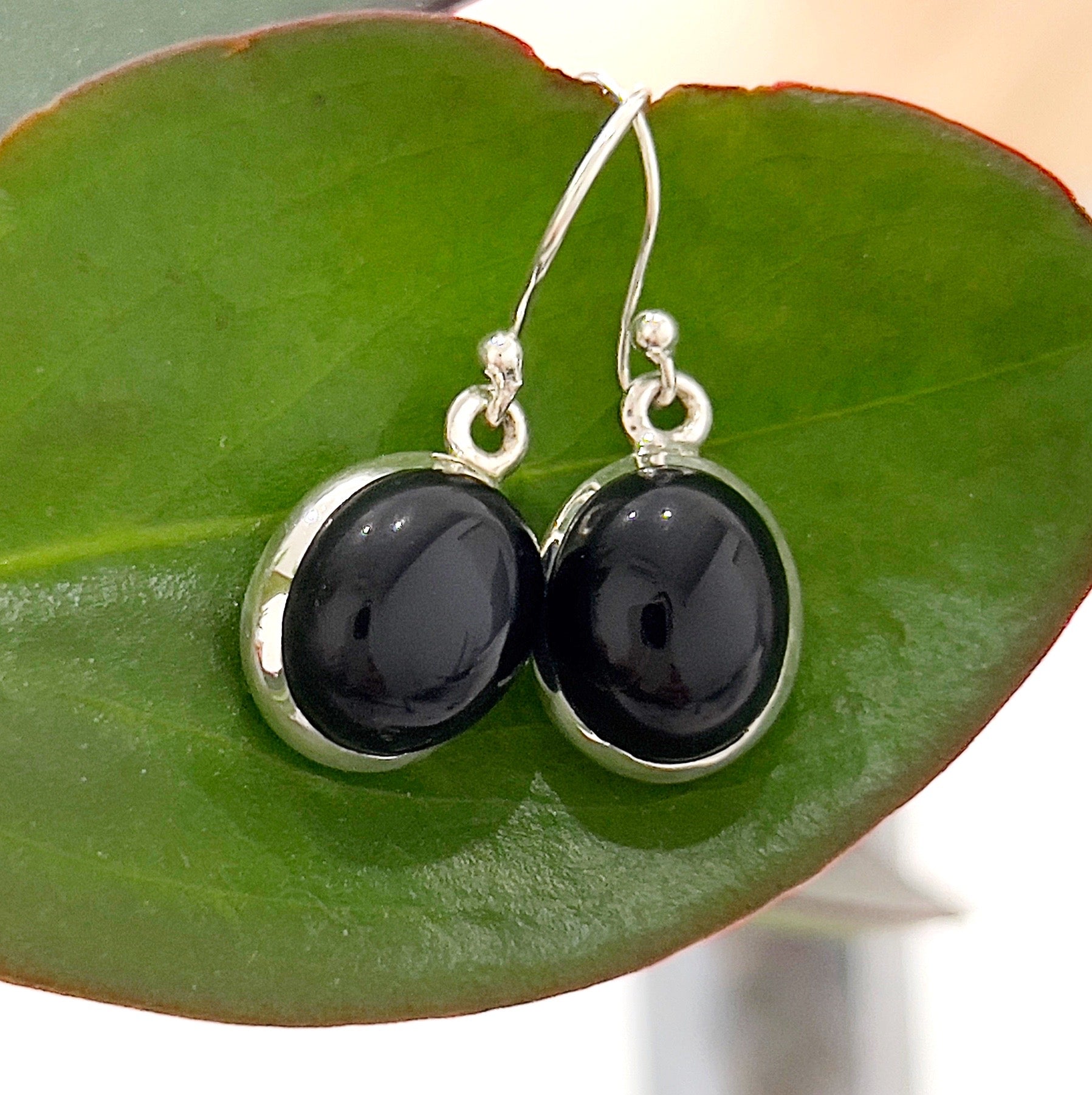 A close-up image of elegant onyx and sterling silver drop earrings, featuring smooth black oval-shaped onyx gemstones set in polished silver frames. The earrings are displayed on a green leaf background, highlighting their sophisticated and minimalist design. Perfect for adding a touch of elegance to any outfit. | Simply Esoteric