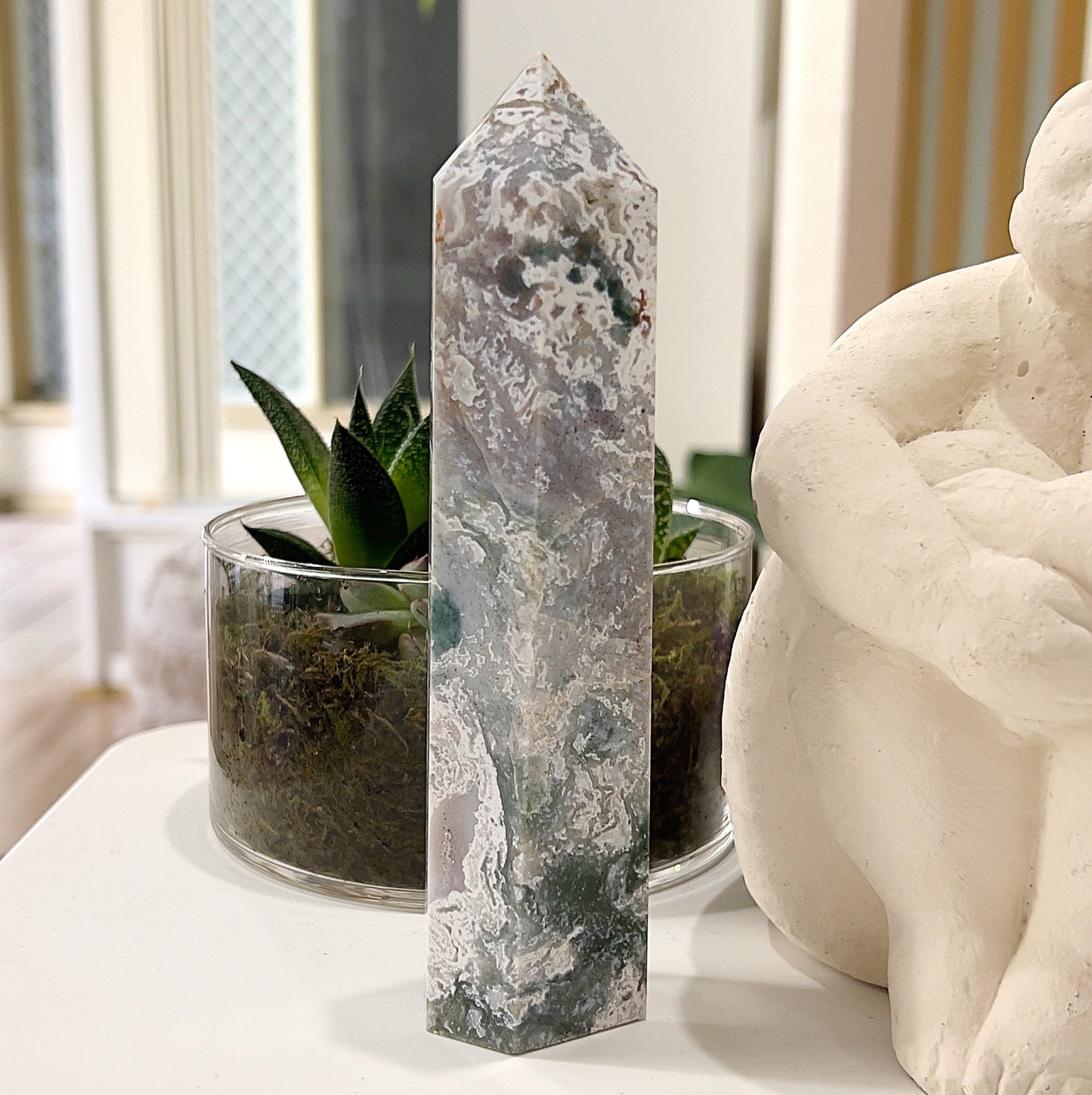 Moss Agate Tower approximately 16cm tall, featuring intricate green, white, and earthy patterns, displayed with a glass pot containing a succulent plant and a white sculpture in the background. | Simply Esoteric