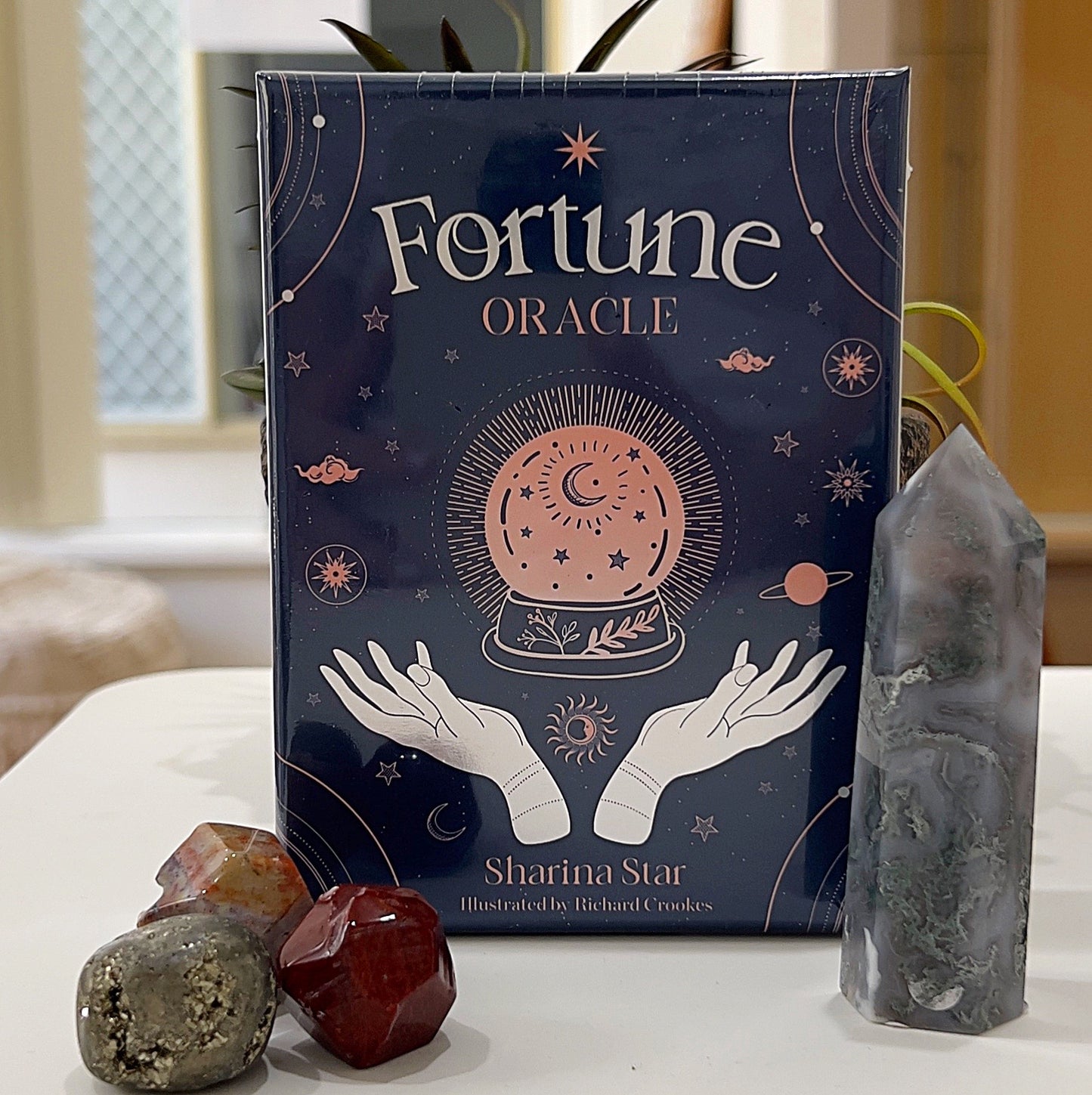 A neatly displayed set of *Fortune Oracle Cards* by Sharina Star, illustrated by Richard Crookes, featuring an elegant dark blue box with celestial-themed artwork, including a crystal ball, stars, planets, and mystical hands. The box is surrounded by polished gemstones and a crystal tower, placed in a serene setting with a light background, creating a mystical and inviting atmosphere. | Simply Esoteric