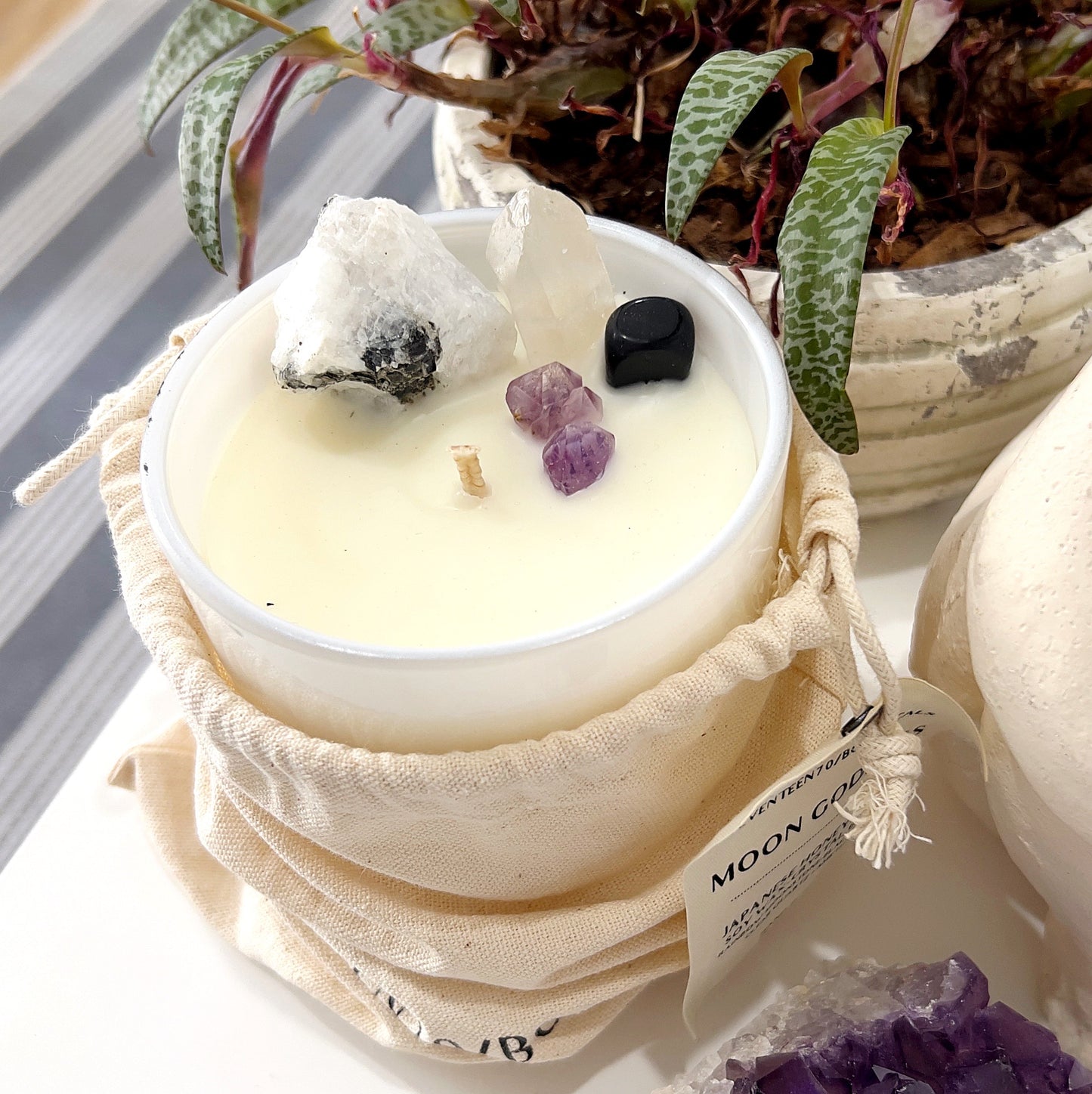 A white candle in a frosted glass container is infused with various crystals, including Rainbow Moonstone, Clear Quartz, Black Obsidian, and Amethyst. The candle is accompanied by a tag that reads "Moon Goddess" and mentions its crystal-infused properties and a 60+ hour burn time. The candle is set against a backdrop of a potted plant and other decorative crystals. The scene evokes a sense of tranquility and spiritual ambiance. | Simply Esoteric