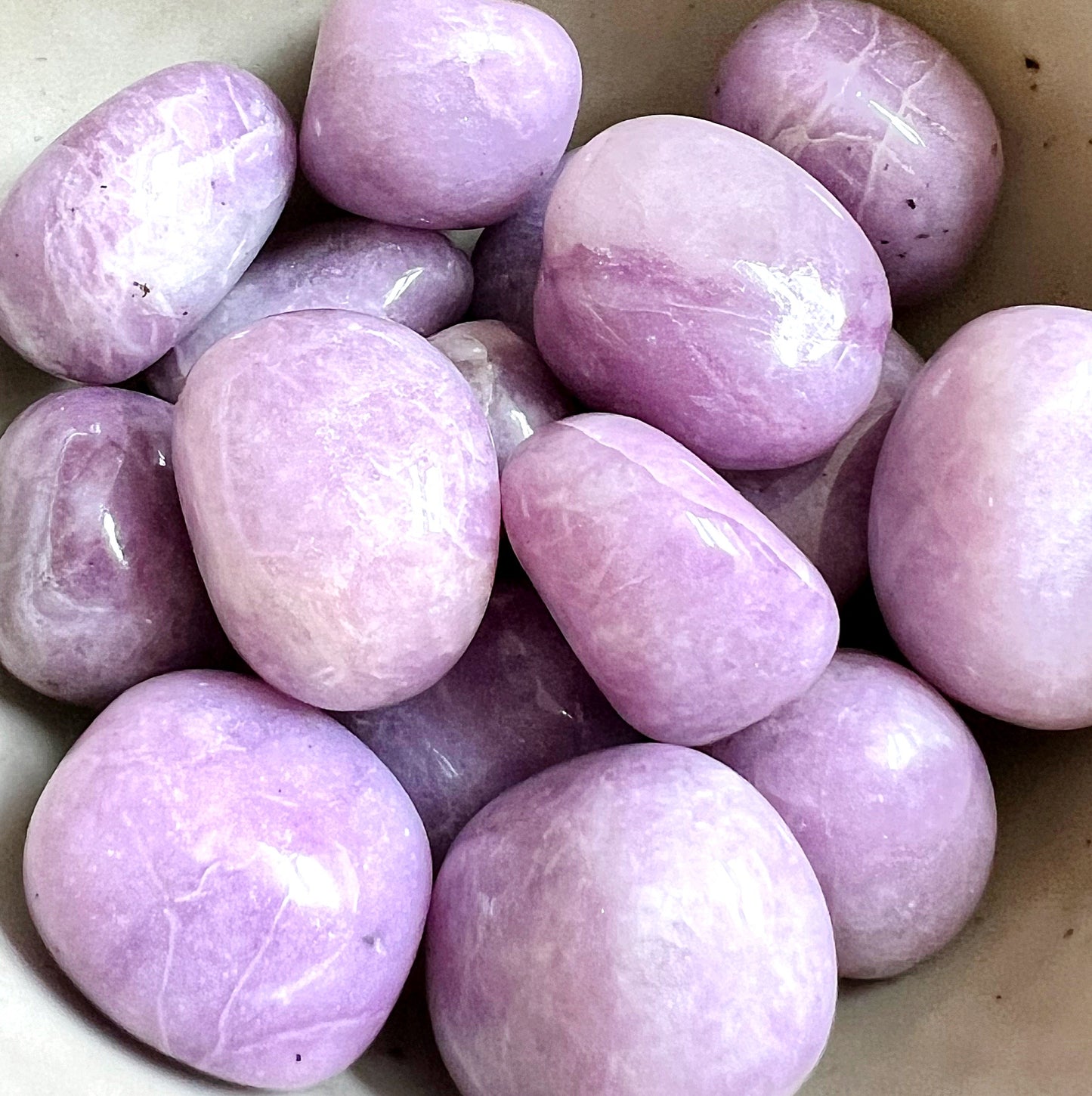 A collection of polished lepidolite tumbled stones displayed in a pile. These stones have a smooth, glossy texture and a rich lavender color with subtle variations in hue and occasional mica inclusions that give them a slight shimmer. Each stone is oval and uniquely patterned, reflecting natural variations typical of lepidolite. | Simply Esoteric