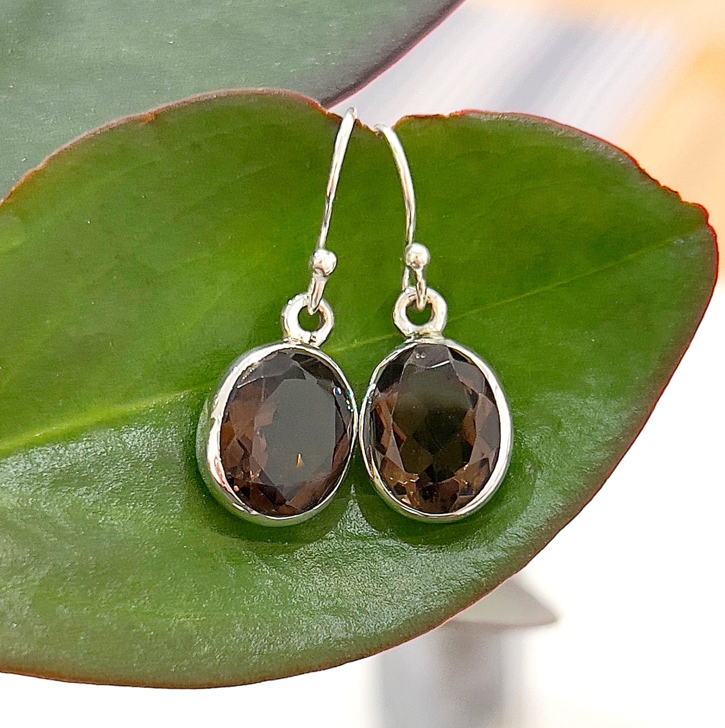 A close-up image of elegant smokey quartz sterling silver drop earrings, featuring oval-cut, rich brown gemstones set in polished sterling silver. The earrings are displayed against a green leaf backdrop, highlighting their classic and sophisticated design. Perfect for adding a touch of natural elegance to any outfit. | Simply Esoteric