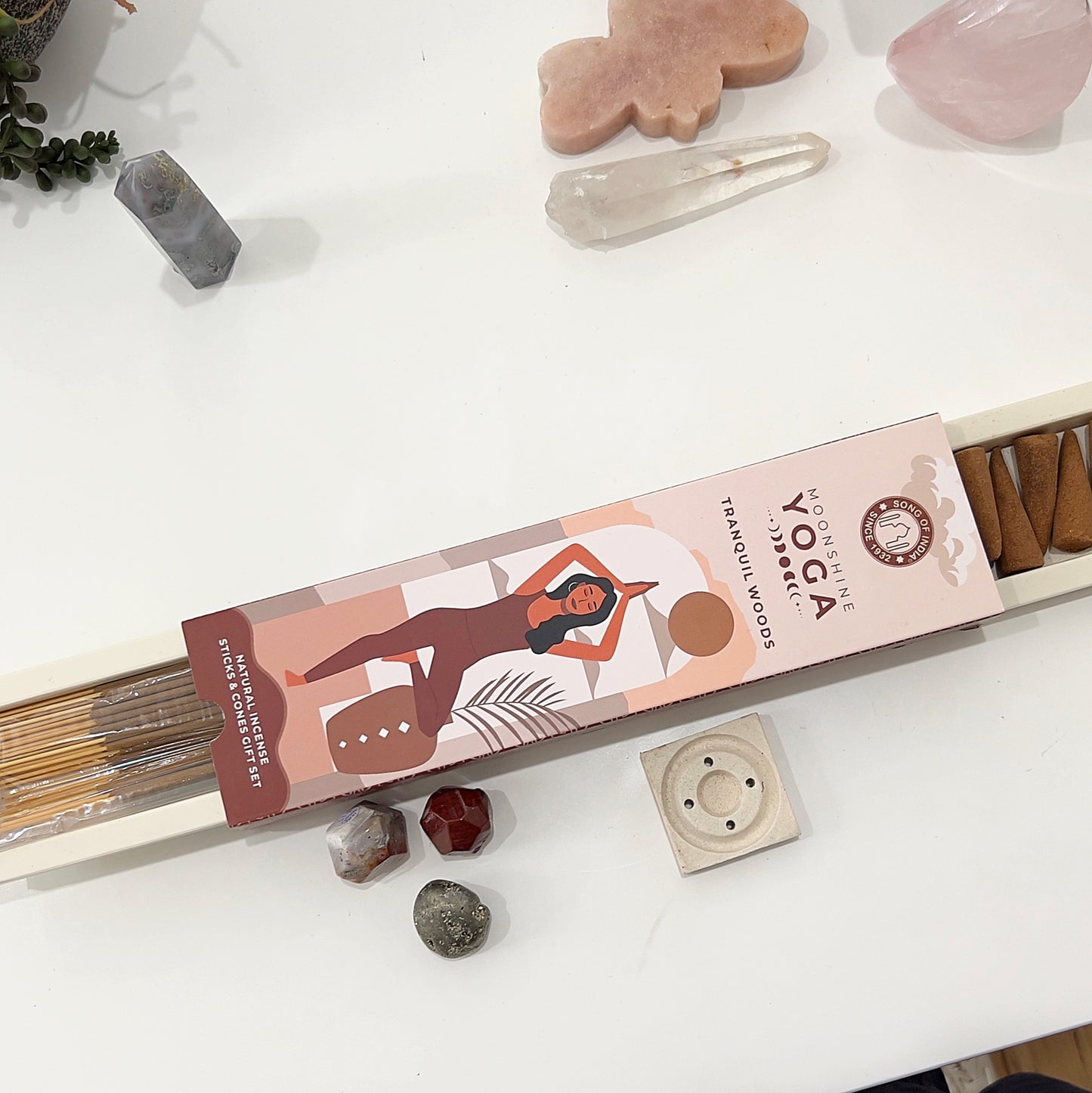 "Moonshine Yoga Tranquil Woods Incense Gift Pack featuring natural incense sticks and cones, along with a stylish incense holder. The outer packaging showcases an illustration of a woman in a yoga pose against a warm earthy background. An open view reveals the incense sticks, cones, and holder, arranged on a white surface surrounded by crystals and decorative plants. | Simply Esoteric
