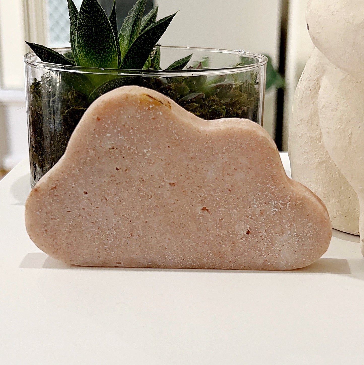 A beautifully crafted Pink Amethyst Cloud, measuring 12cm by 7cm, displayed on a white surface with a background of green plants. The cloud-shaped crystal features a soft pink hue, symbolizing love and emotional healing, with a polished surface highlighting its natural beauty and intricate details.