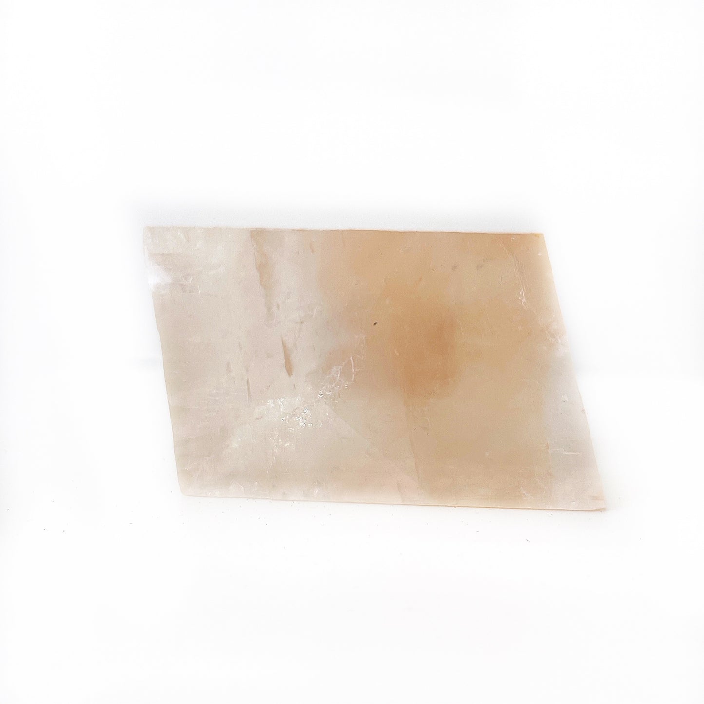 A rectangular piece of Optical Orange Calcite with translucent, warm peachy-orange tones, resting diagonally on a white surface. The smooth, polished stone displays natural lines and cloud-like inclusions, creating a soft, ethereal appearance. | Simply Esoteric