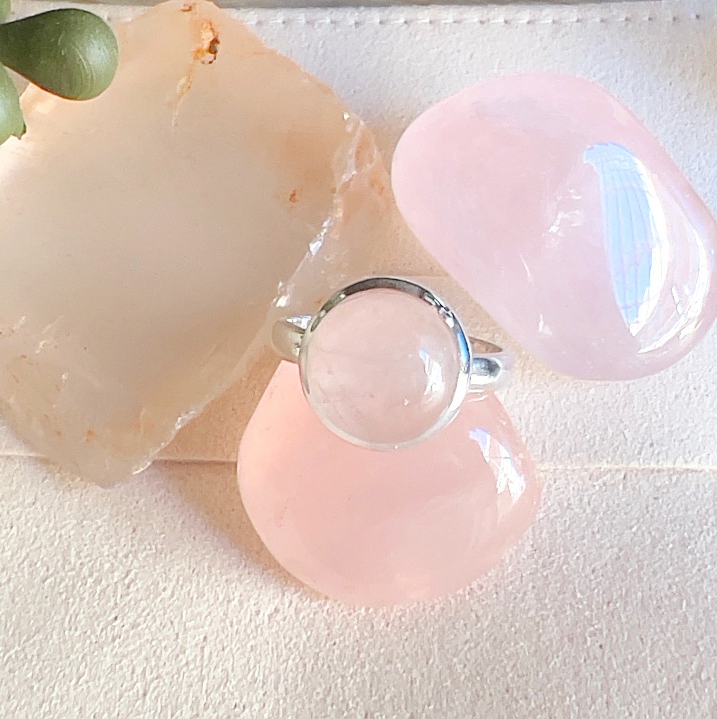 Sterling silver Rose Quartz ring with a round soft pink stone, displayed on a light surface with rose quartz crystals surrounding it. | Simply Esoteric