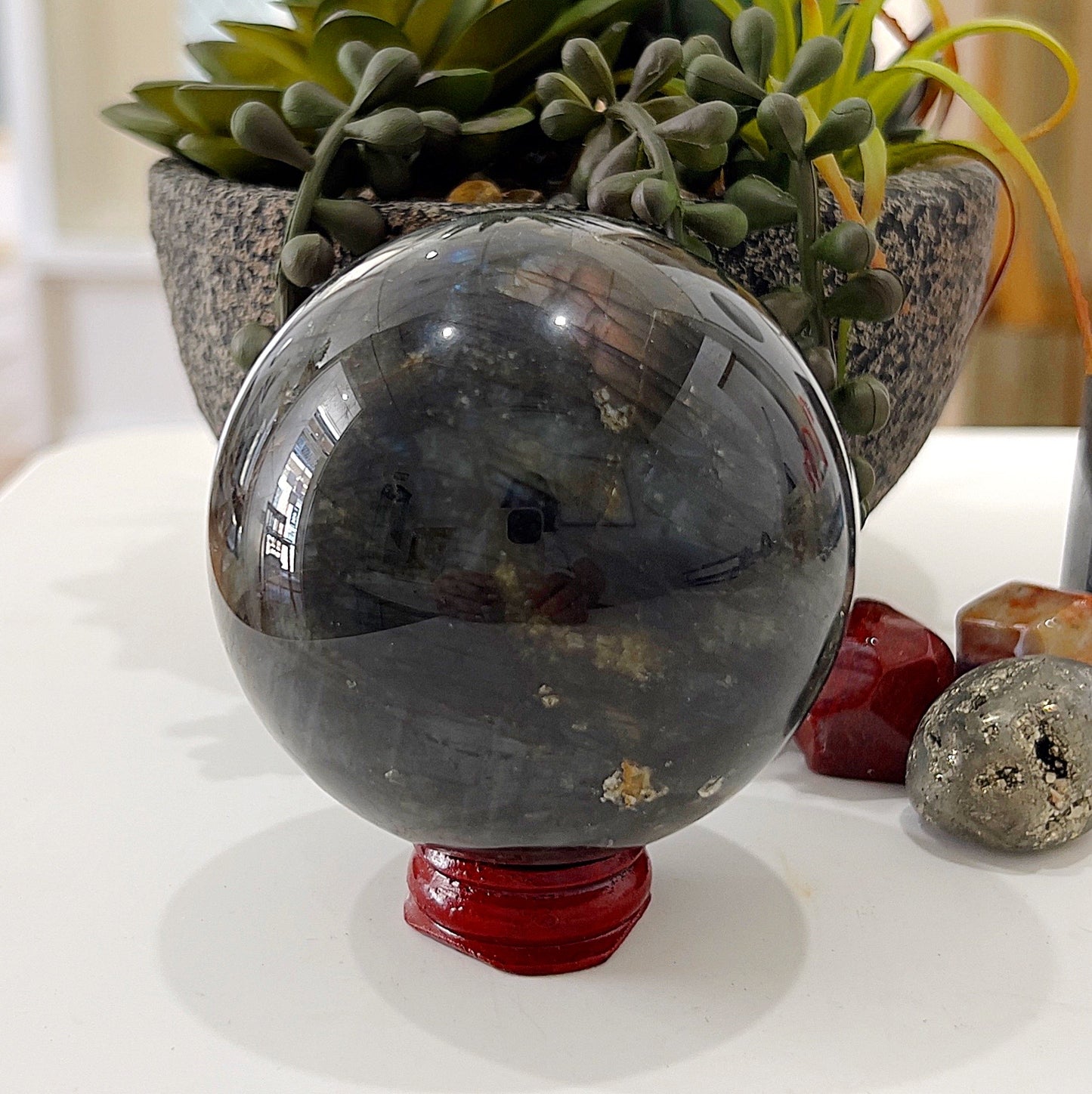 A polished Labradorite sphere approximately 7cm in diameter, resting on a red stand. The sphere features a dark base with subtle flashes of iridescent blue, green, and gold hues, placed in front of a backdrop of vibrant green succulents for an earthy and mystical display. | Simply Esoteric
