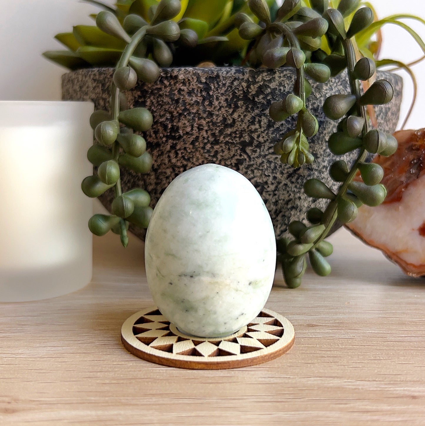 A polished jadeite egg approximately 5cm tall, featuring soft pale green tones with gentle white marbling. Displayed upright on a wooden geometric base, set against a natural backdrop with succulents, a candle, and agate slice for a calming, earthy presentation. | Simply Esoteric