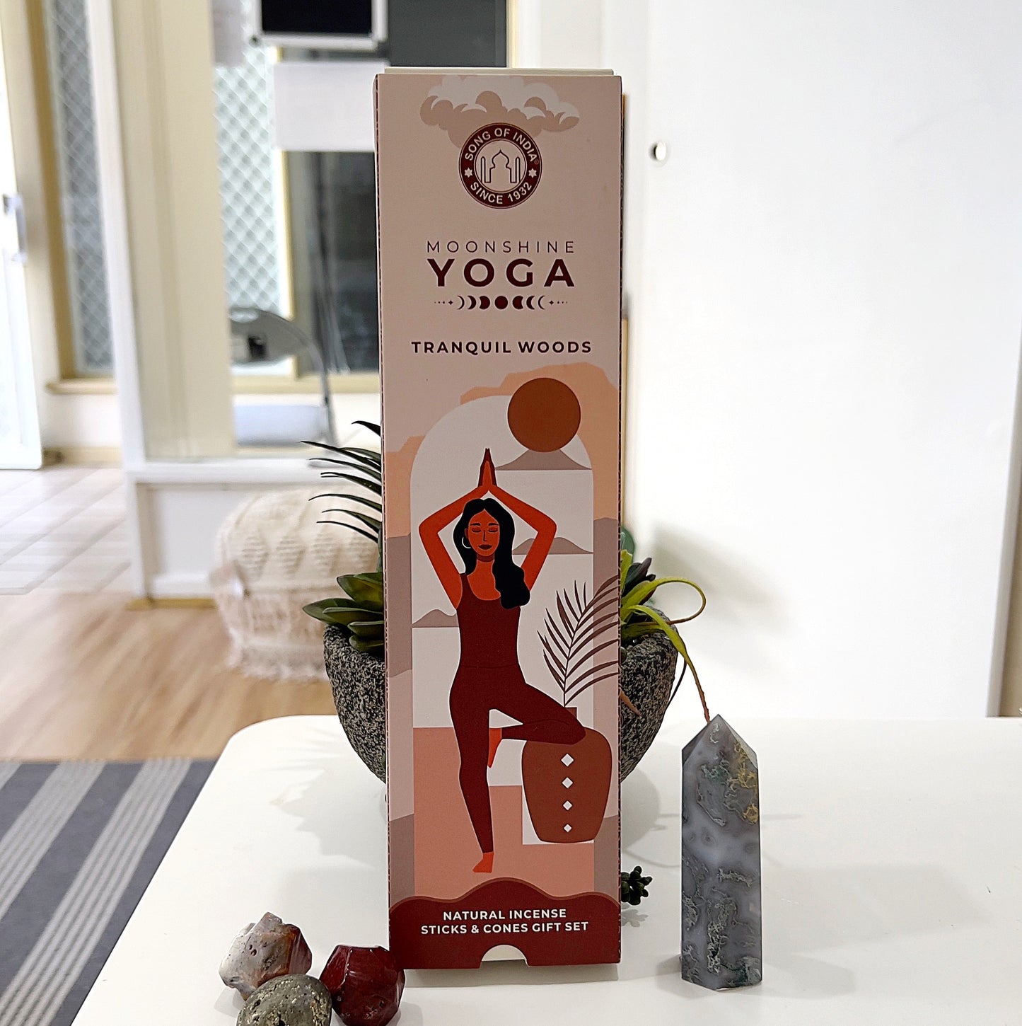 "Moonshine Yoga Tranquil Woods Incense Gift Pack featuring natural incense sticks and cones, along with a stylish incense holder. The outer packaging showcases an illustration of a woman in a yoga pose against a warm earthy background. An open view reveals the incense sticks, cones, and holder, arranged on a white surface surrounded by crystals and decorative plants. | Simply Esoteric