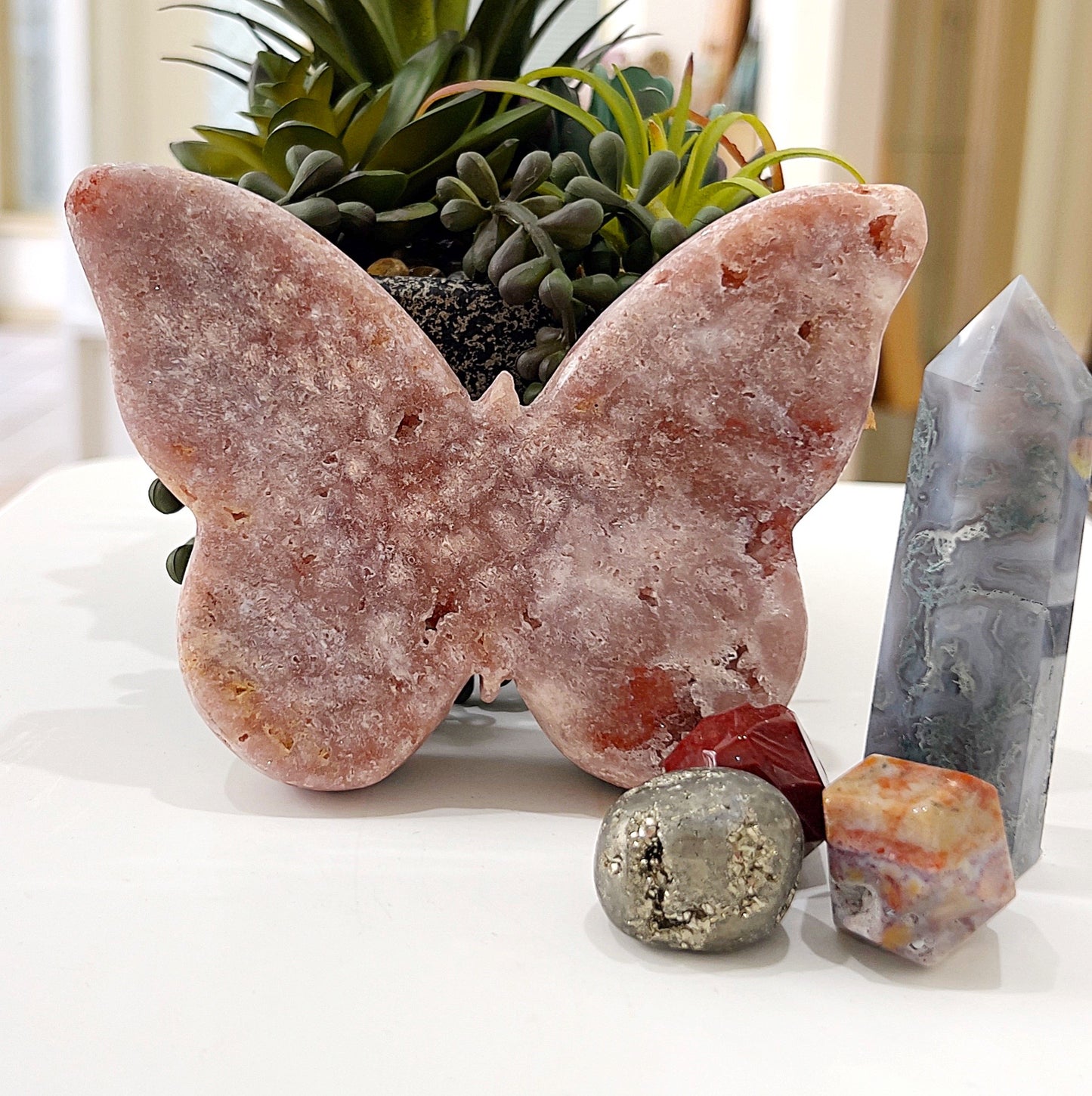 A pink amethyst butterfly sculpture approximately 14cm wide, showcasing soft blush hues and intricate crystal formations, placed on a white surface surrounded by decorative stones and succulents for a serene and natural aesthetic. | Simply Esoteric