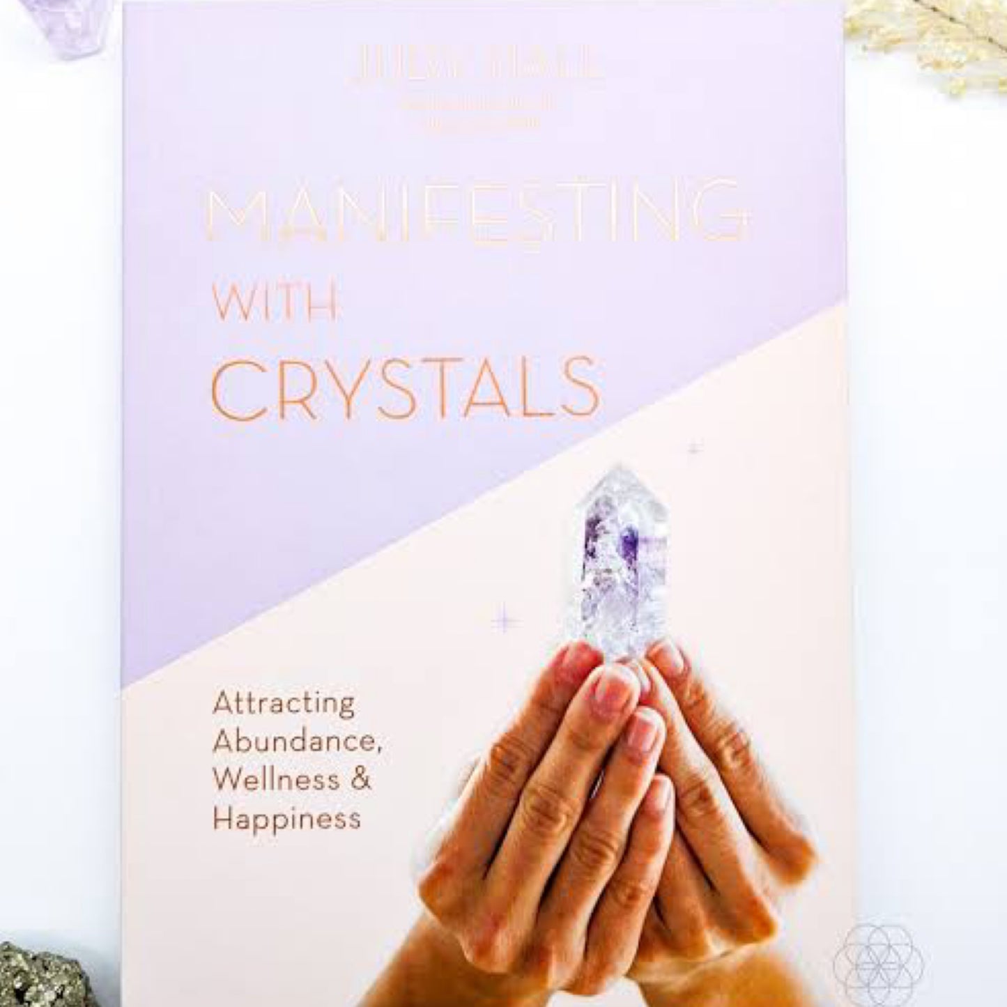 'Manifesting With Crystals' by Judy Hall, featuring an image of a hand holding a radiant, multifaceted crystal, symbolizing the power of crystals in aiding personal manifestation. The title is prominently displayed in a clear, elegant typeface against a peaceful background that invites readers to unlock the secrets of crystal energy for their aspirations. | Simply Esoteric