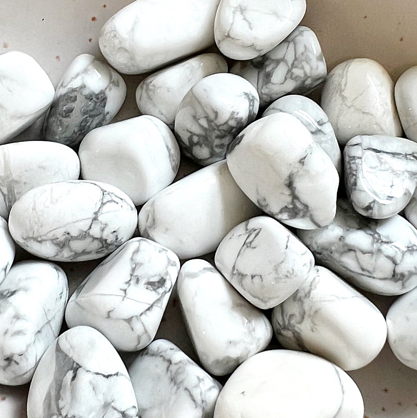 A close-up image of multiple white howlite tumbled stones with distinctive grey veining. The stones have a smooth, polished surface and are irregularly oval-shaped, showcasing the natural marbled patterns of white and grey that characterize howlite. The soft, muted tones of the stones provide a serene and calming visual. | Simply Esoteric