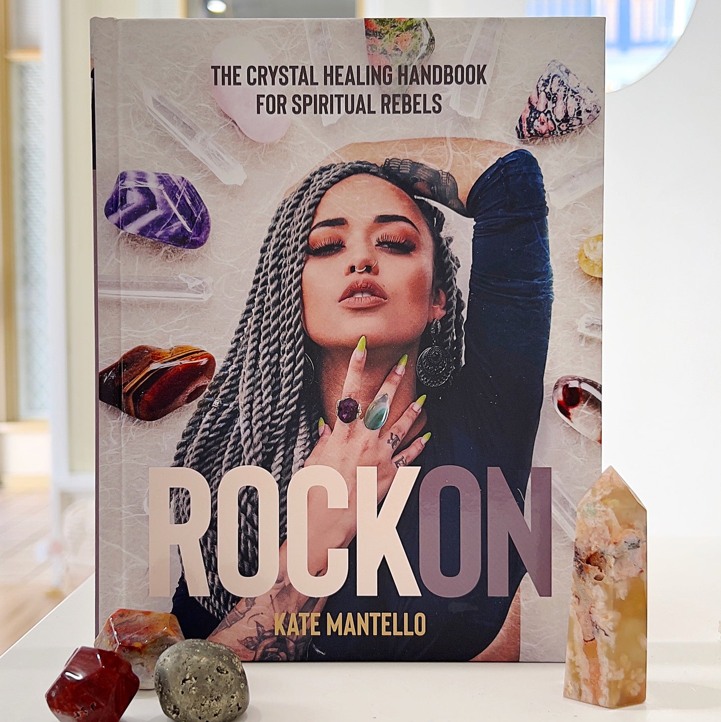 A bold and vibrant book cover for "Rock On: The Crystal Healing Handbook for Spiritual Rebels" by Kate Mantello. The cover features a confident woman with long braided hair, striking makeup, and vibrant accessories, including crystal rings. Surrounded by a dynamic array of colorful crystals, the design conveys a modern and edgy approach to spirituality. Additional crystals and gemstones are placed in the foreground for added depth and aesthetic appeal. | Simply Esoteric