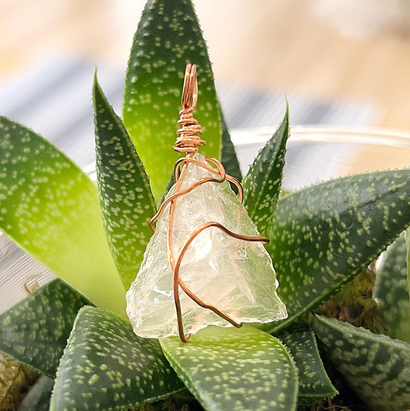 A close-up image of a rose quartz wire wrap pendant in rose gold colour, set against a background of green succulents. The soft pink rose quartz crystal is encased in delicate rose gold-coloured wire, showcasing its natural beauty and elegance. | Simply Esoteric