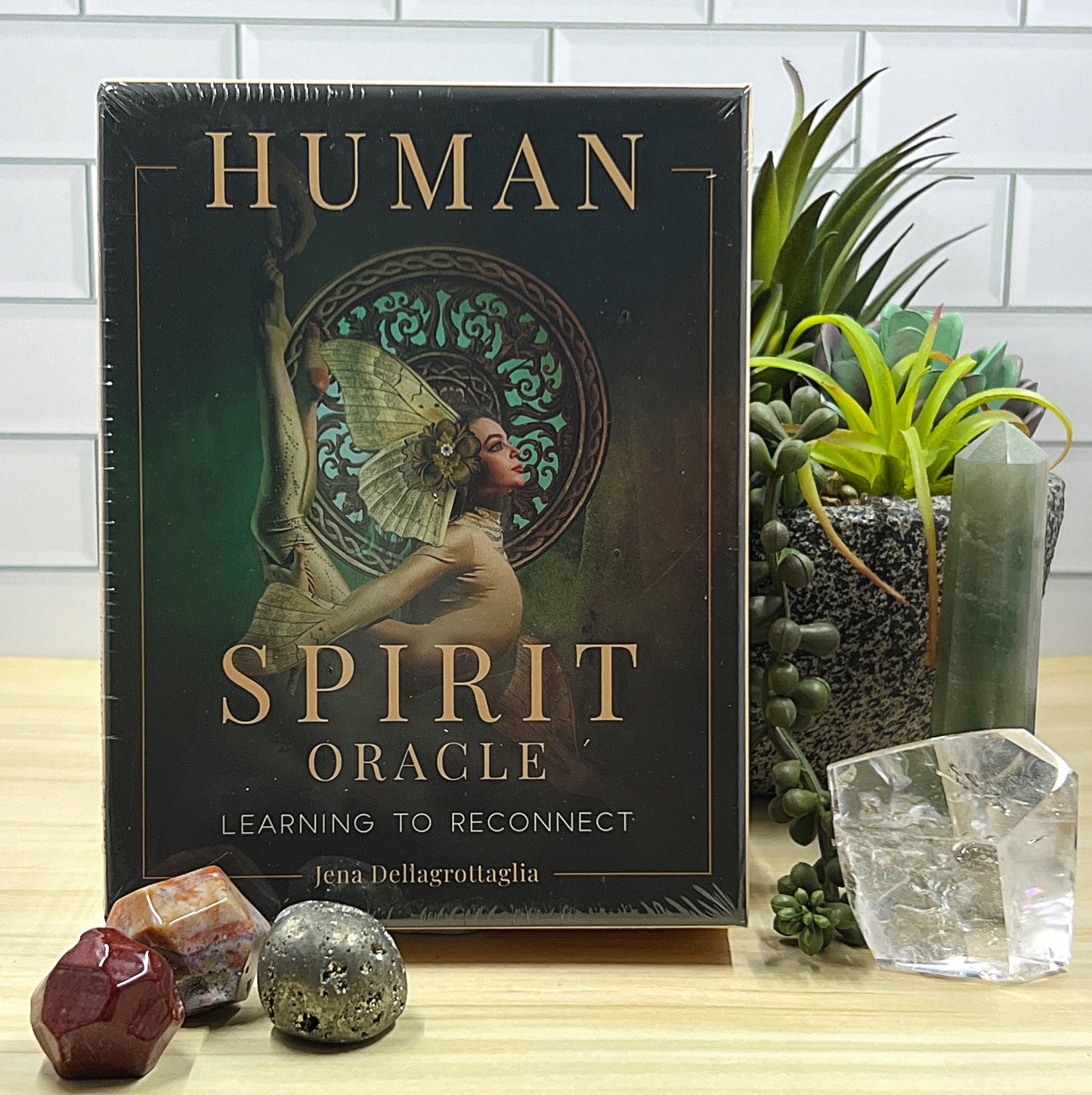 Human Spirit Oracle Cards by Jena Dellagrottaglia - a 44-card deck with intricate artwork displayed on the front of the box, featuring a woman with butterfly-like wings and an ornate circular backdrop. The box is set against a natural display with crystals, a plant, and gemstones for a serene and mystical aesthetic. | Simply Esoteric