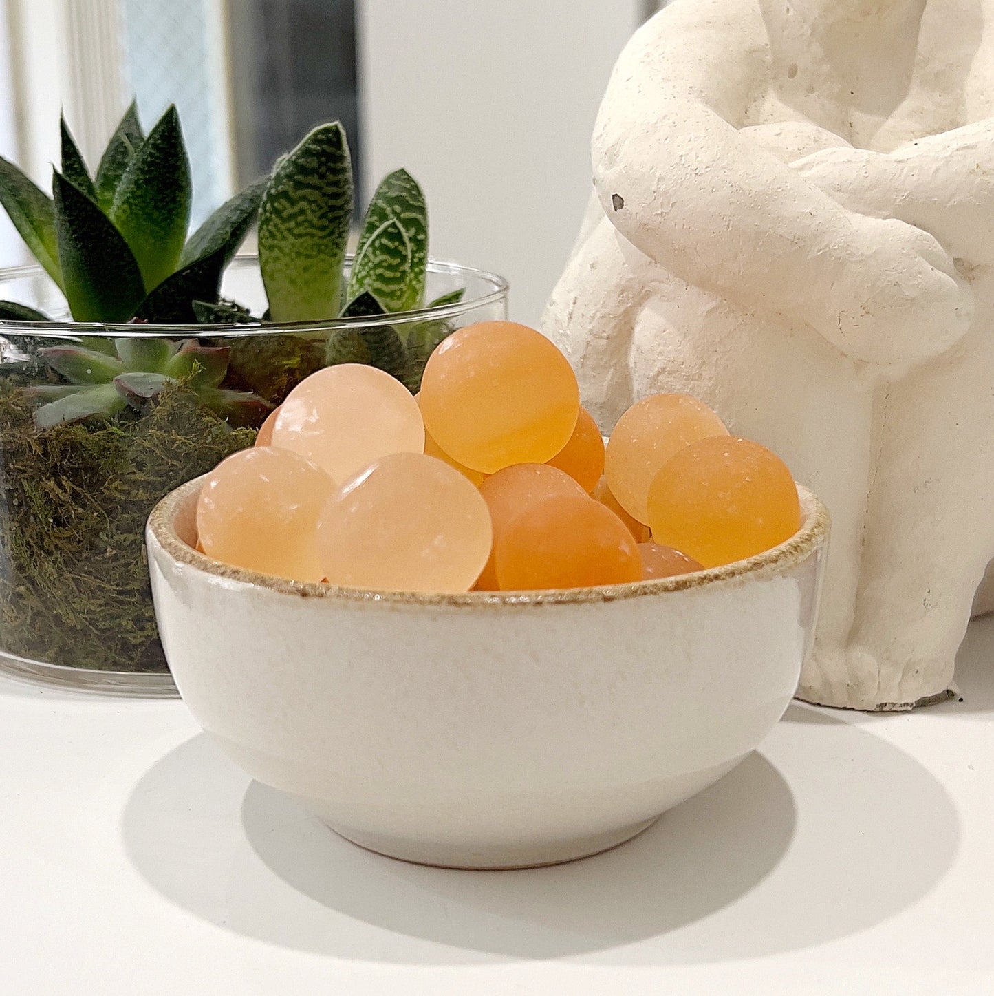 A bowl of polished orange selenite balls, each approximately 2.5cm in diameter, displaying a warm, peachy-orange hue. The bowl is set next to a small potted plant and a white decorative statue, creating a serene and elegant scene. | Simply Esoteric