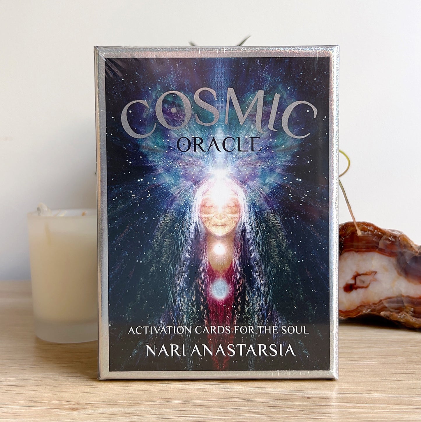 A boxed set of “Cosmic Oracle: Activation Cards for the Soul” by Nari Anastarsia, featuring ethereal artwork of a radiant cosmic being with glowing chakras. Displayed on a wooden surface with a candle, plant, and agate slice in the background, creating a serene and spiritual setting. | Simply Esoteric