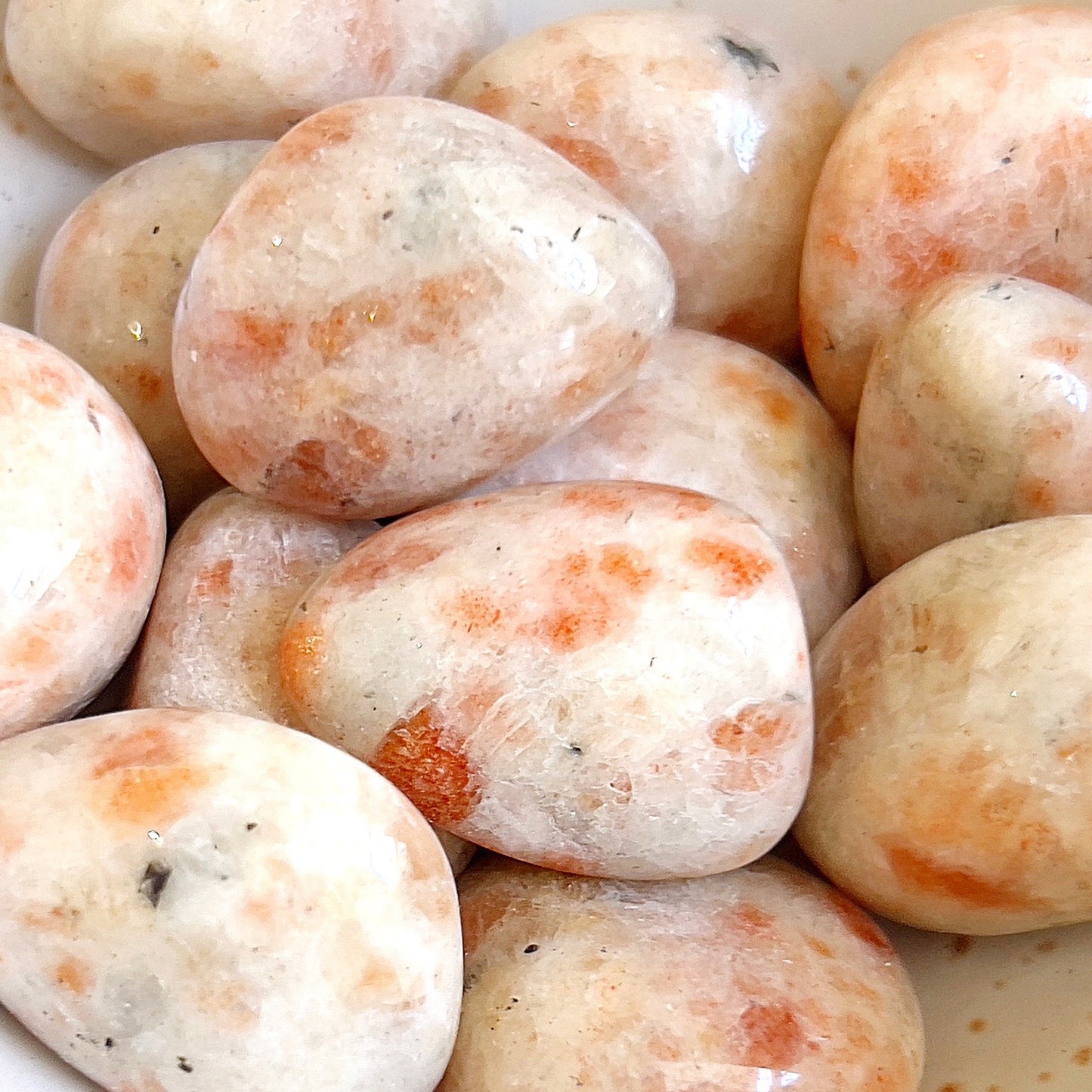 A collection of polished sunstone tumbled stones with a smooth finish and an interplay of orange and cream hues. Some stones exhibit natural sparkling inclusions and flecks of darker mineral deposits, adding to their unique charm. The stones vary slightly in shape and size, with a warm and inviting appearance reminiscent of a sunlit day. | Simply Esoteric