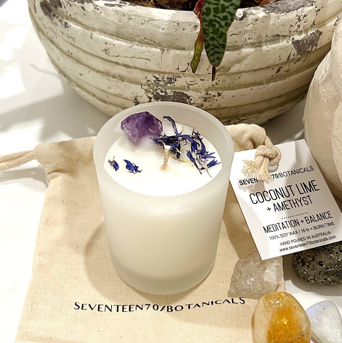 A crystal-infused candle from Seventeen70 Botanicals, featuring a blend of coconut and lime scents, adorned with an Amethyst gemstone. The white soy wax candle, displayed in a simple glass container, emits a fresh, tropical aroma, accented with floral notes and placed beside a small canvas bag and various crystals. Ideal for meditation and promoting balance.