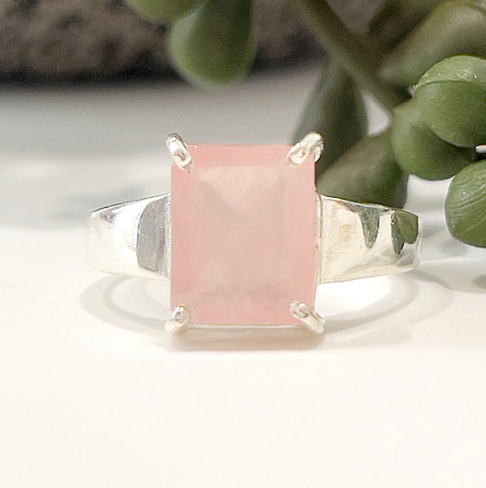 A close-up image of a sterling silver ring featuring a rectangular-cut rose quartz gemstone in a prong setting. The soft pink hue of the stone contrasts beautifully with the polished silver band. The ring is displayed on a white surface with green succulent plants in the background, highlighting its elegant and natural aesthetic. | Simply Esoteric