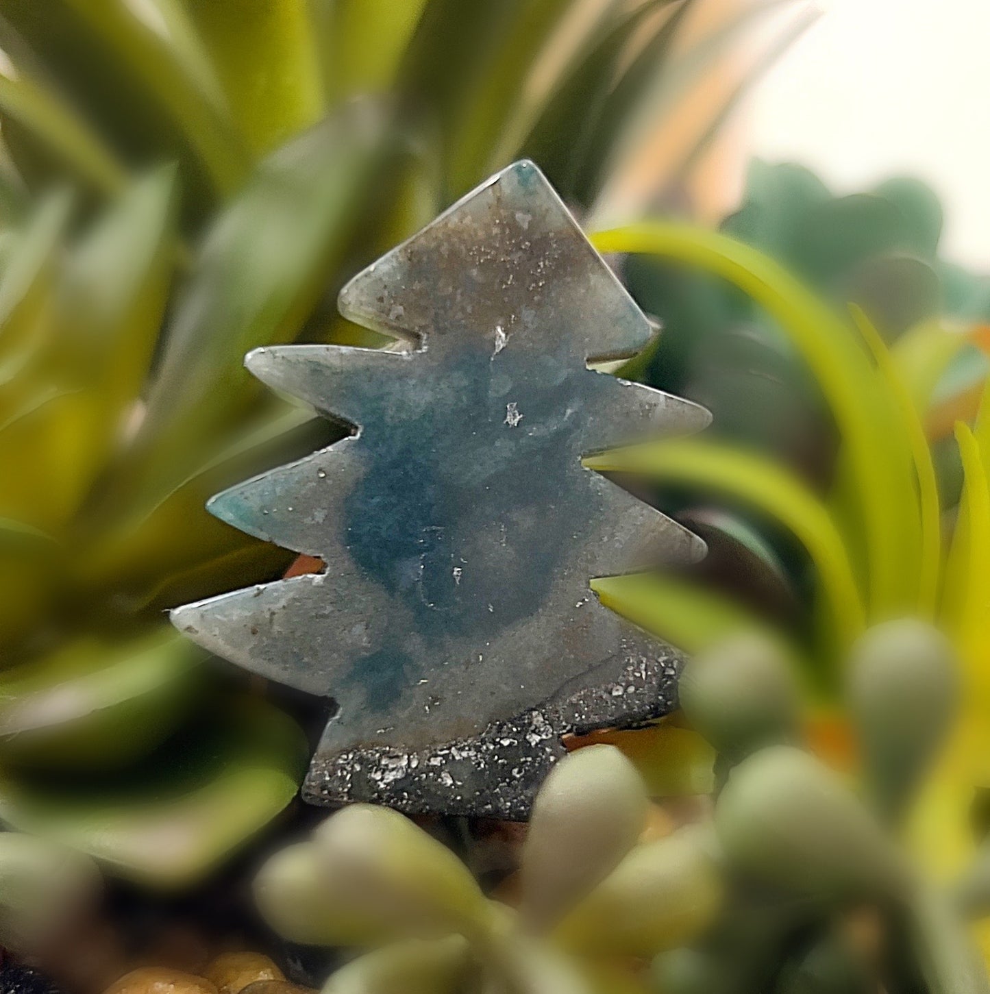 A small moss agate tree carving, approximately 4cm tall, with soothing green hues and natural patterns, placed in front of a background of green succulents. The carving resembles the shape of a pine tree, showcasing the earthy, grounding energy of the moss agate stone. | Simply Esoteric