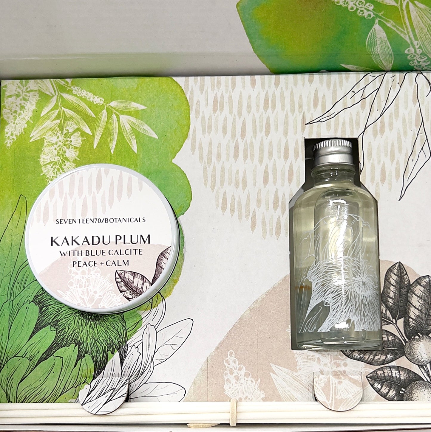 A beautifully arranged Kakadu Plum Reed Diffuser Gift Set by Seventeen70 Botanicals, featuring a soy wax candle with a 20+ hour burn time and a reed diffuser. The set is presented in a decorative box with a green and white botanical theme, showcasing the candle in a tin labeled "Kakadu Plum with Blue Calcite Peace + Calm" alongside a bottle of diffuser oil. The overall aesthetic is clean and inviting, emphasizing the natural and calming elements of the product. | Simply Esoteric