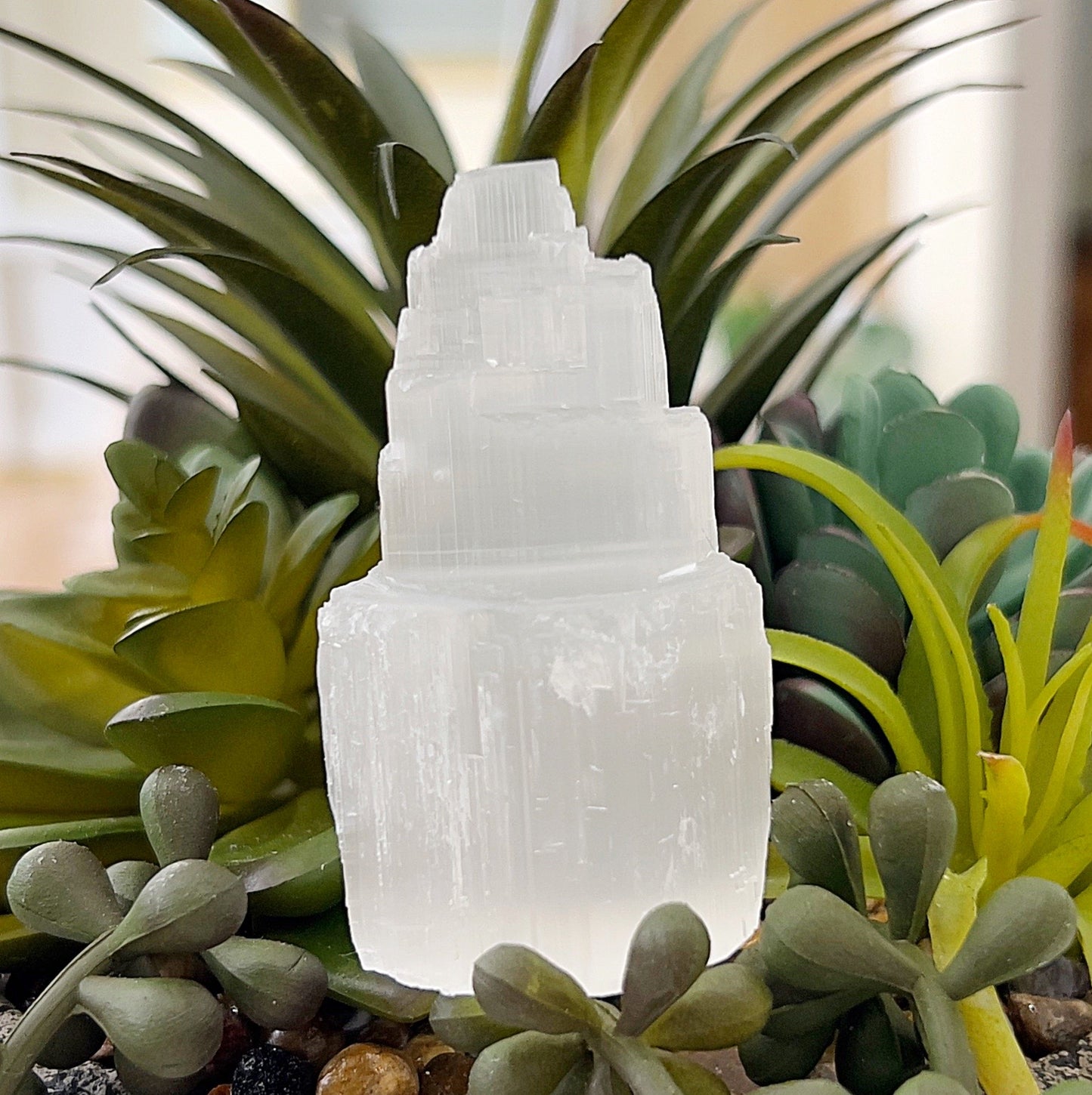 A 6cm raw selenite tower with a stepped, natural design, displayed against a background of vibrant green succulents. The tower’s translucent white surface reflects soft light, highlighting its layered texture. Perfect for energy cleansing and meditation spaces. | Simply Esoteric