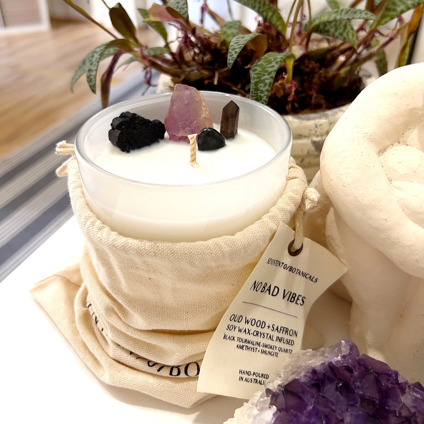 A soy wax candle labeled "No Bad Vibes" by Seventeen70 Botanicals, featuring a crystal-infused top with Black Tourmaline, Shungite, Smokey Quartz, and Amethyst. The candle is in a frosted glass container, accompanied by a natural cotton drawstring bag. The label indicates the candle has a rich and woody fragrance of Oud and Saffron, and is vegan-friendly with a 60+ hour burn time. In the background, there are plants and other crystals, enhancing the serene and protective vibe of the setup. | Simply Esoteric
