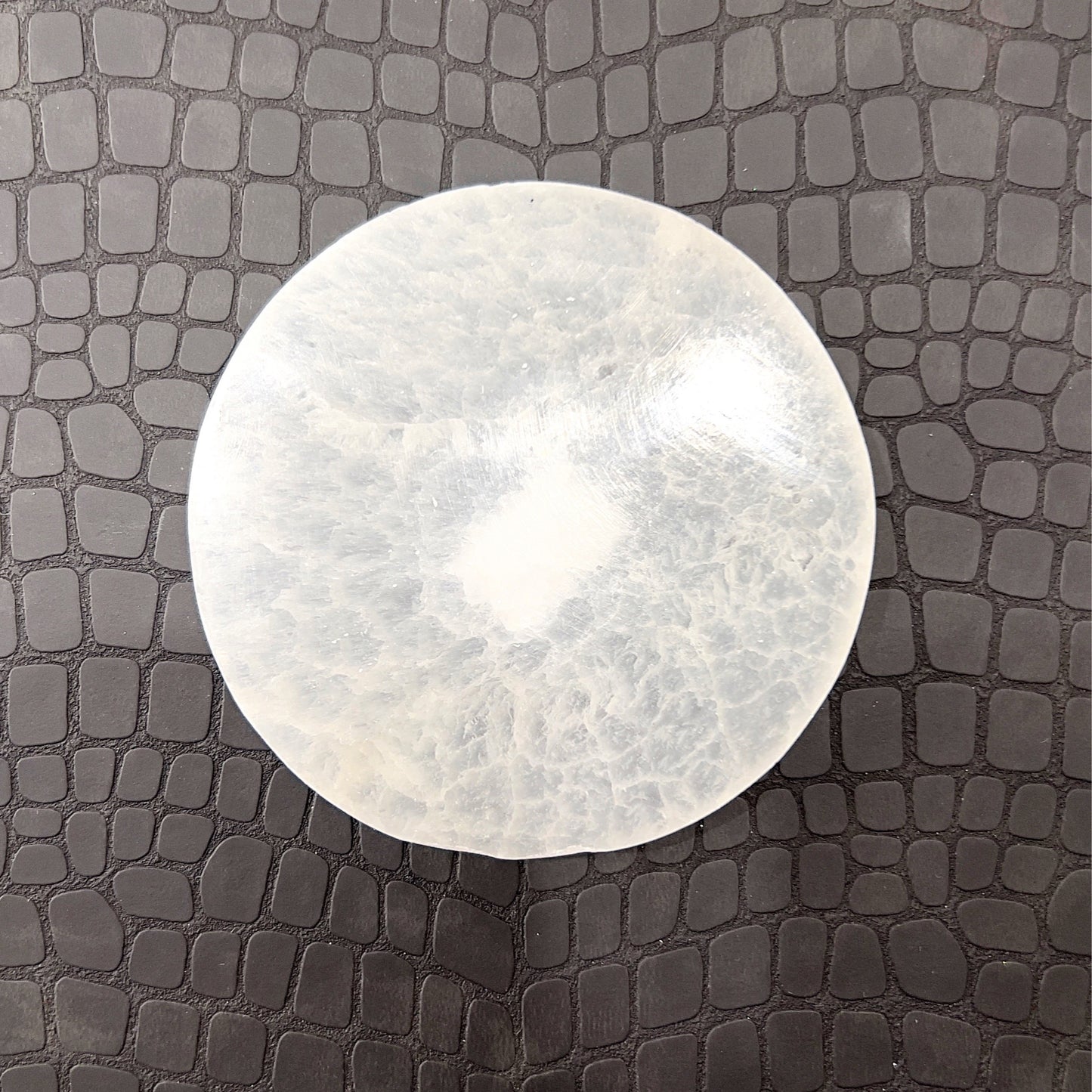 A polished selenite charging plate with a smooth, oval shape and a pearly white glow. Known for its energy-cleansing properties, the plate has a flat surface ideal for placing other crystals upon for purification and recharging, highlighted against a simple background that accentuates its pure, clean appearance. | Simply Esoteric