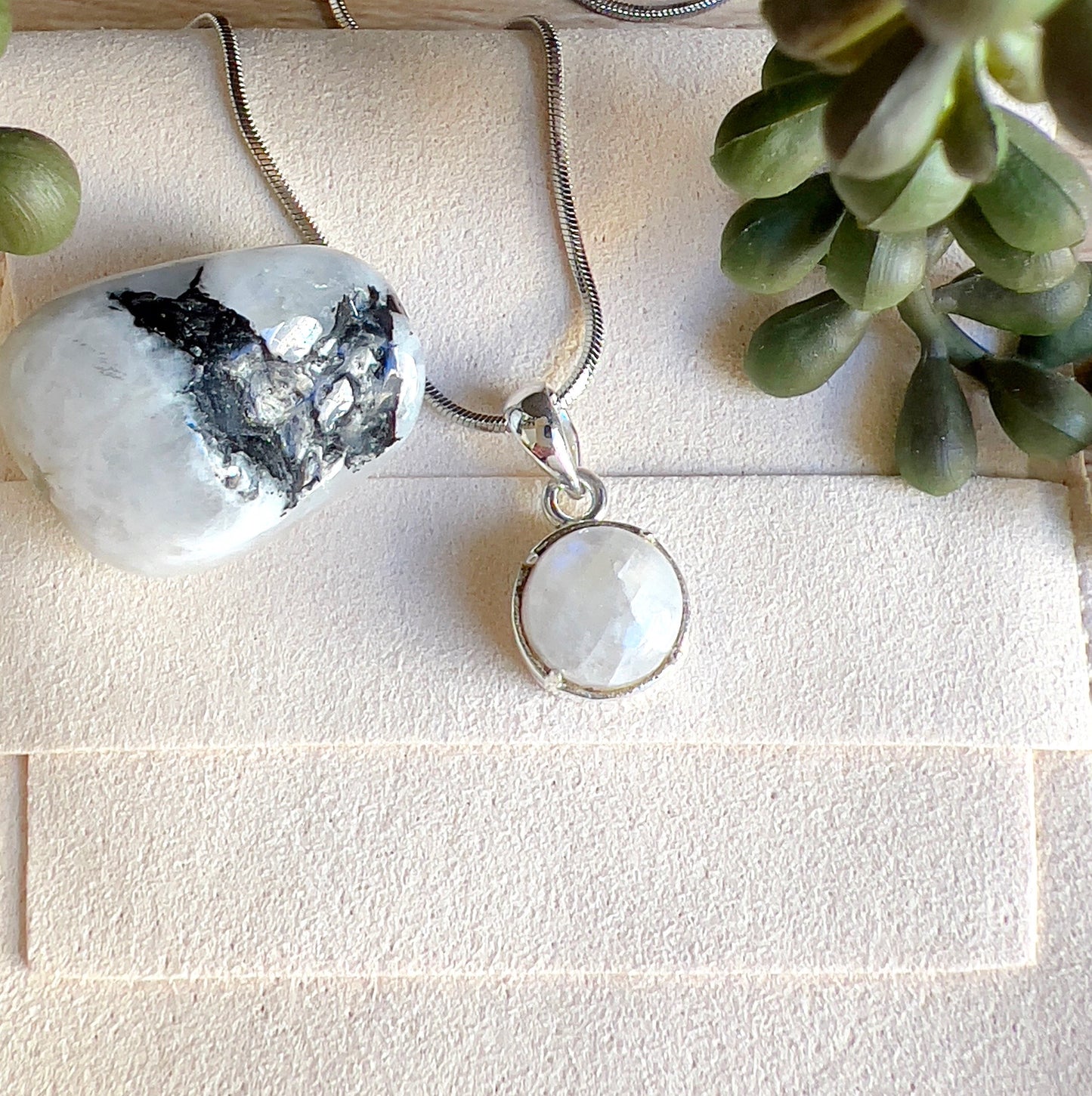 Sterling silver Moonstone pendant with a luminous round stone, displayed on a silver chain alongside polished Moonstone crystals on a light surface. | Simply Esoteric
