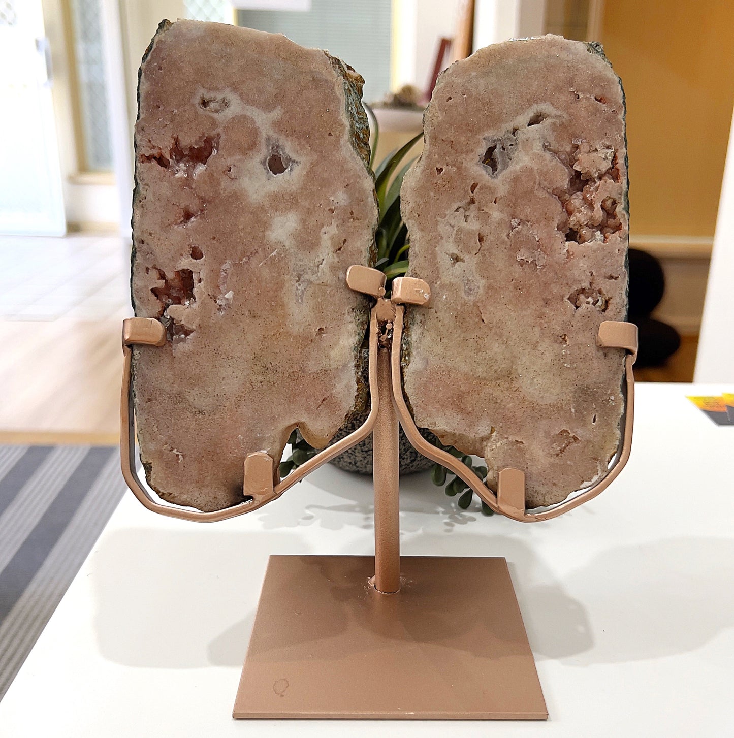 A pair of pink amethyst slices displayed on a stylish rose gold metal stand. The slices feature soft blush tones with natural crystal formations and patterns, adding an elegant and calming touch. The stand securely holds the slices upright, making it ideal for home decor or spiritual spaces. The background includes subtle greenery for a natural aesthetic. | Simply Esoteric