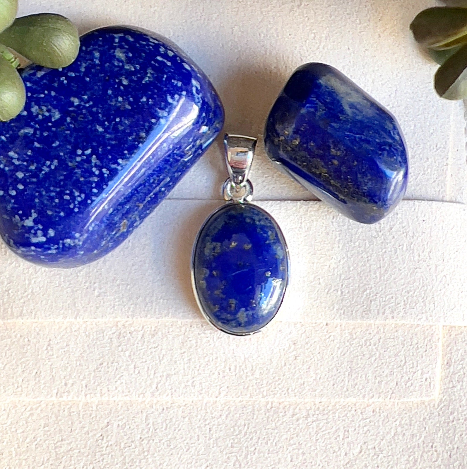 Sterling silver Lapis Lazuli pendant with oval blue stone featuring natural golden flecks, displayed alongside polished Lapis Lazuli tumbles on a light surface. | Simply Esoteric