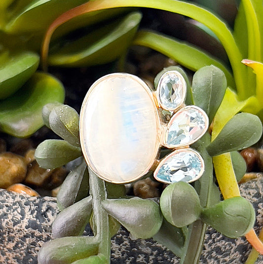**Alt Text:** A sterling silver adjustable ring featuring a luminous oval moonstone at the center, surrounded by three faceted blue topaz gemstones set in a teardrop design, placed against green succulent leaves. The moonstone has a soft, iridescent glow, while the blue topaz stones sparkle with light blue hues. | Simply Esoteric
