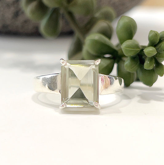 A close-up image of a green amethyst sterling silver ring, size 9, featuring a radiant rectangular-cut green amethyst gemstone in a prong setting. The ring rests on a white surface, accompanied by a small green succulent in the background, highlighting the gemstone's soft green hue and elegant sterling silver band. | Simply Esoteric