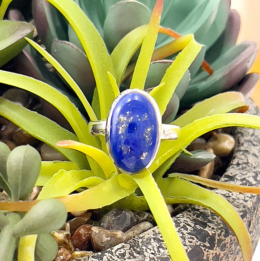 A sterling silver ring featuring an oval-shaped lapis lazuli gemstone, bezel-set with deep blue tones and golden pyrite flecks. The ring rests against a backdrop of vibrant green succulent leaves and natural stones, highlighting its elegant and timeless design. Perfectly crafted in size 7 for a minimalist and refined aesthetic. | Simply Esoteric