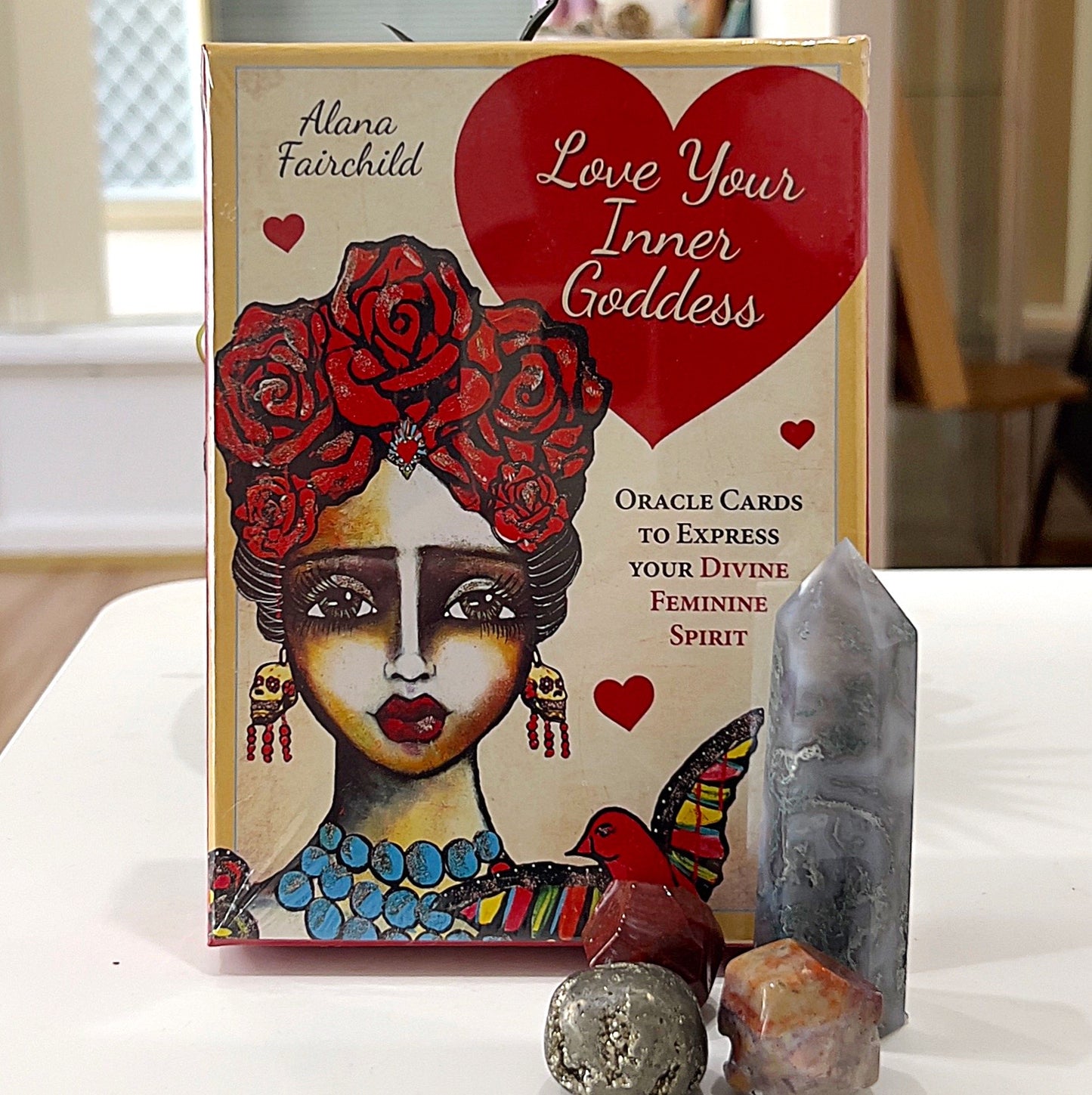 A vibrant image showcasing the *Love Your Inner Goddess Oracle Cards* by Alana Fairchild. The box features stunning artwork of a goddess adorned with red roses and vibrant accessories, symbolising divine feminine energy. Surrounding the box are decorative crystals, including a polished grey tower and sparkling stones, adding to the mystical and spiritual vibe of the scene. | Simply Esoteric