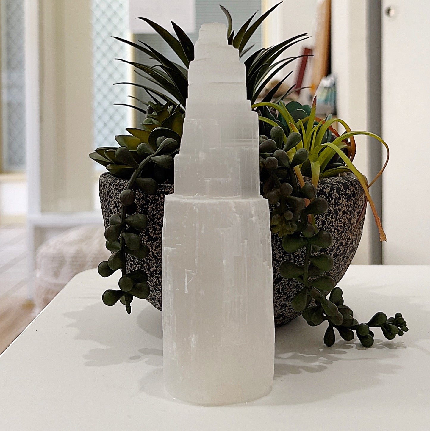 A raw selenite tower approximately 15cm tall, displayed on a table with a background of lush green succulents in a textured grey pot. The tower features a natural, translucent white texture and a tiered design, symbolizing spiritual ascension. Its luminous glow enhances the serene and elegant aesthetic of the scene. | Simply Esoteric