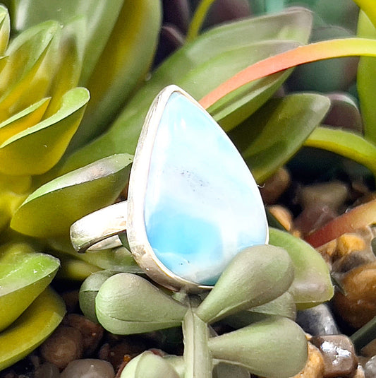 A close-up image of a pear-shaped Larimar gemstone ring set in sterling silver, displayed against a natural background of green succulents and small decorative stones. The ring features soft blue hues with white swirls, representing the serene beauty of the sea, and is designed with an adjustable band for a comfortable fit. | Simply Esoteric