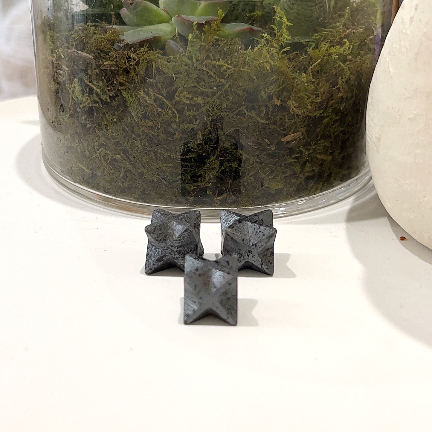 Set of Hematite Merkaba Stars, each approximately 1.5-2cm in size, featuring a dark metallic sheen, displayed with a plant in the background. | Simply Esoteric