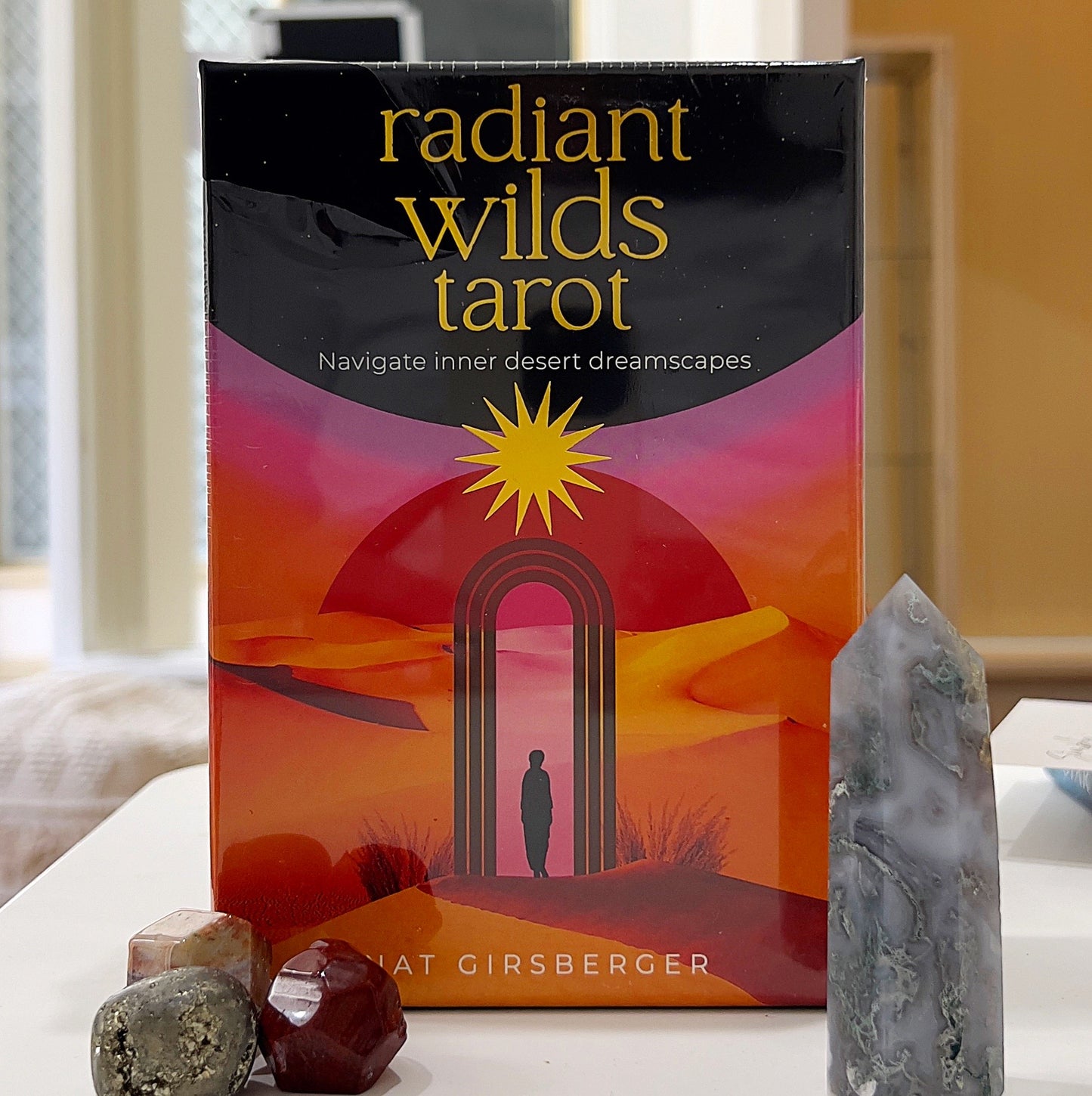 A photo of the Radiant Wilds Tarot deck by Nat Girsberger, featuring a vibrant cover with a desert landscape, a glowing archway, and a silhouette of a person standing beneath a radiant star. The tarot box is surrounded by natural crystal pieces, including a polished gemstone and a quartz tower, placed on a light surface with a soft, neutral background. | Simply Esoteric