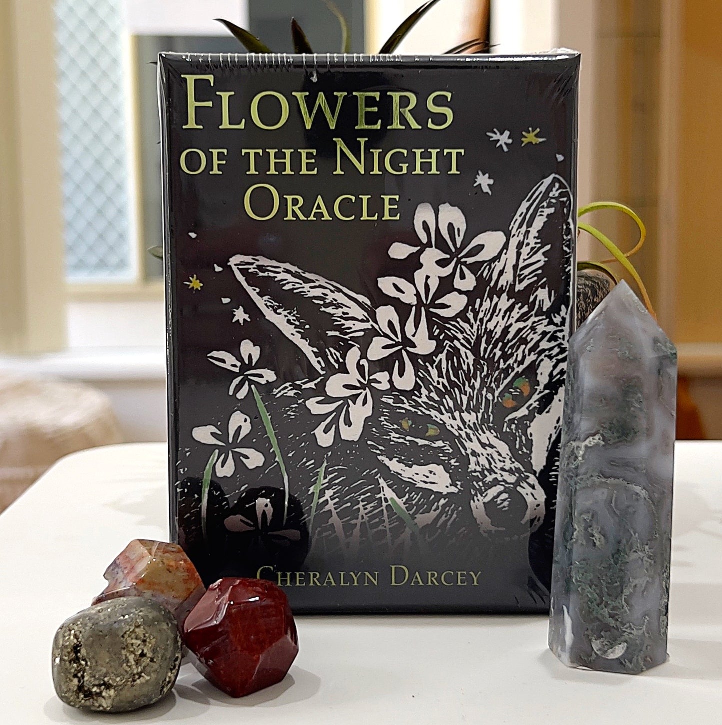 A close-up image of the *Flowers of the Night Oracle* card deck by Cheralyn Darcey. The packaging features an elegant black design with a fox illustration surrounded by white flowers, evoking a mystical, nighttime theme. The deck is displayed upright, accompanied by crystal accents, including red and silver gemstones, and a gray tower crystal in the background. This setup highlights the deck's nature-inspired and spiritual essence. | Simply Esoteric