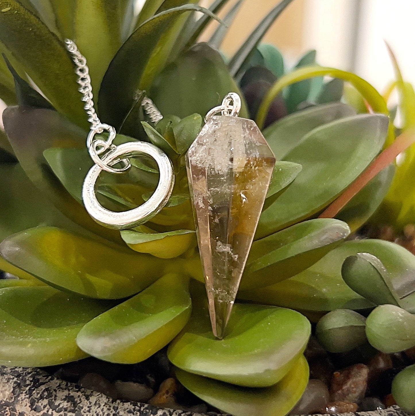 A close-up image of a smoky quartz pendulum with a faceted crystal point. The pendulum hangs from a silver-tone chain featuring a sleek circular ring for handling. The crystal's earthy brown hues shimmer with natural variations, resting against a backdrop of vibrant green succulent plants, creating a serene and grounding aesthetic. | Simply Esoteric