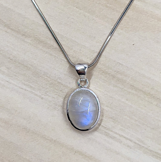 A stunning Moonstone Sterling Silver Pendant featuring an oval-shaped moonstone with a soft blue glow, bezel-set in polished sterling silver. The pendant is displayed on a wooden surface with a delicate chain loop, emphasizing its elegant and timeless design. Chain not included. | Simply Esoteric
